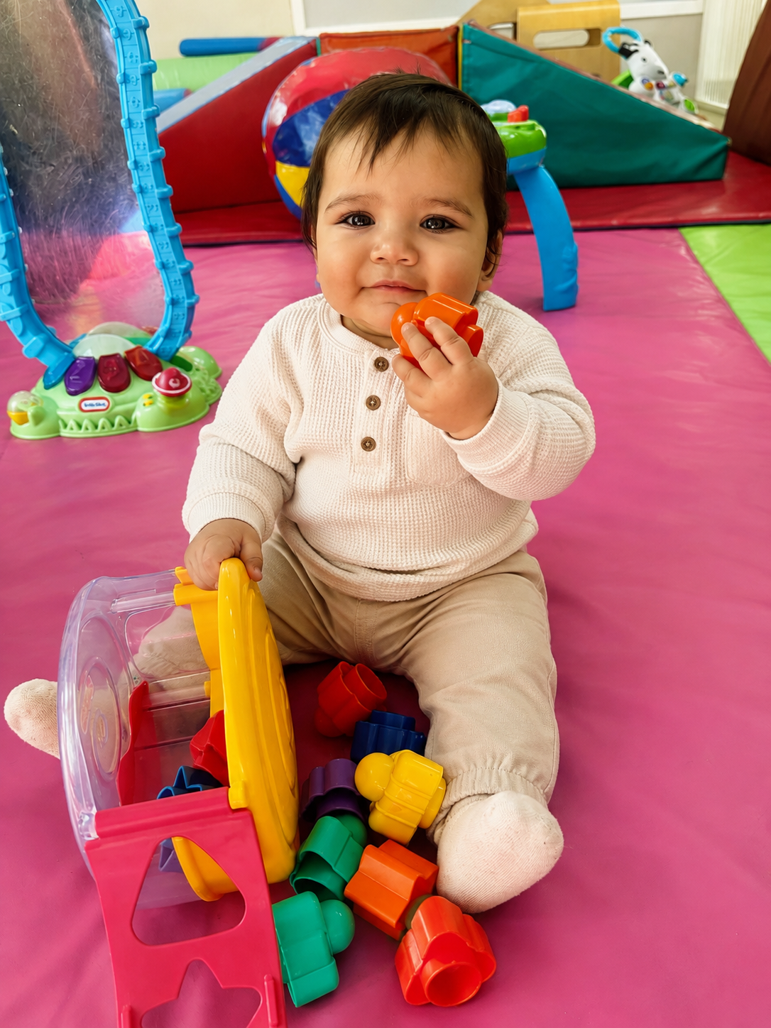 A young child sitting on a colorful play mat holding and playing with toy building blocks, surrounded by more blocks and toys in a playroom.