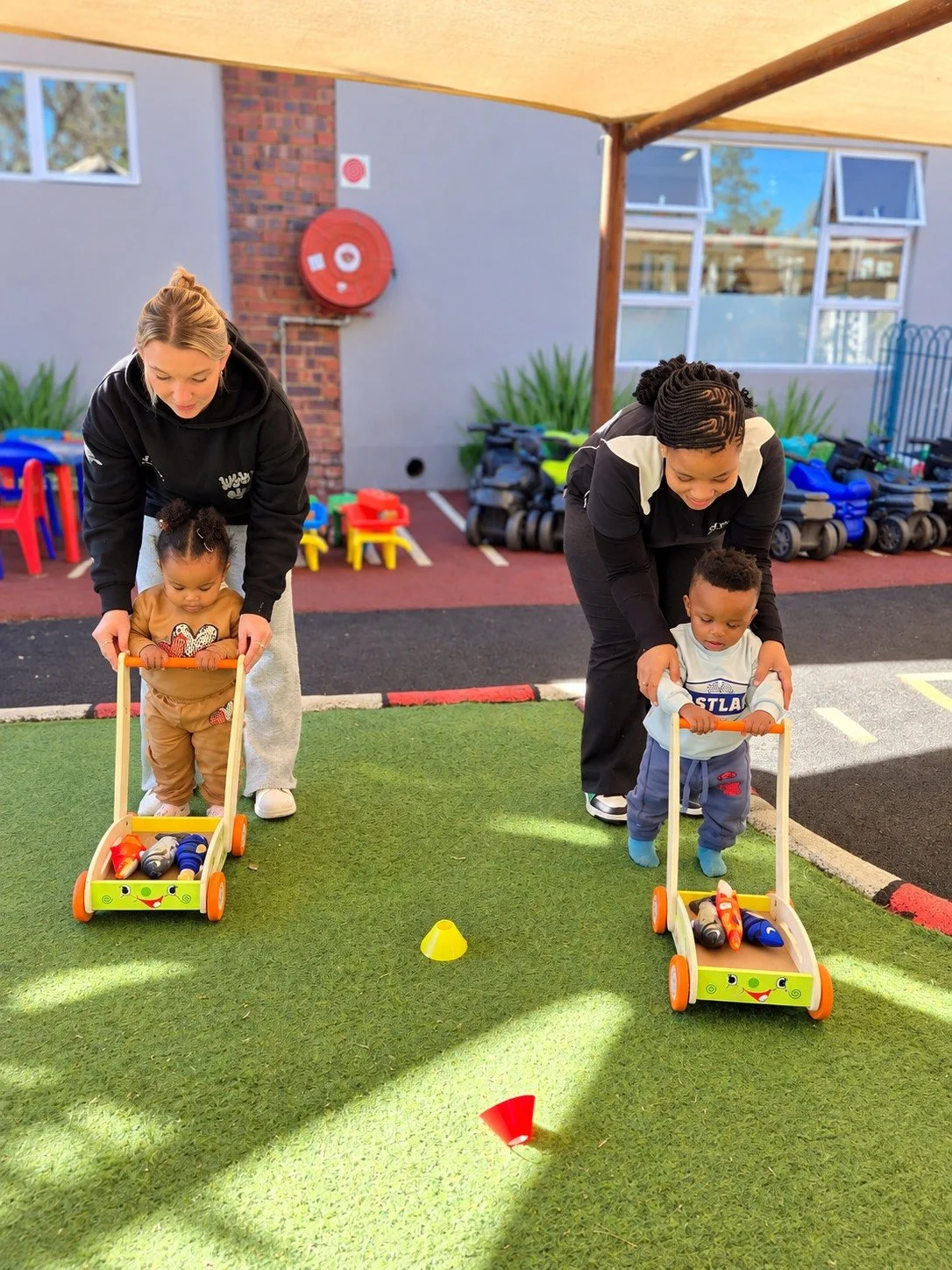 Two young children in toddler wheelbarrows being guided by two adult women during an outdoor activity at a playground.