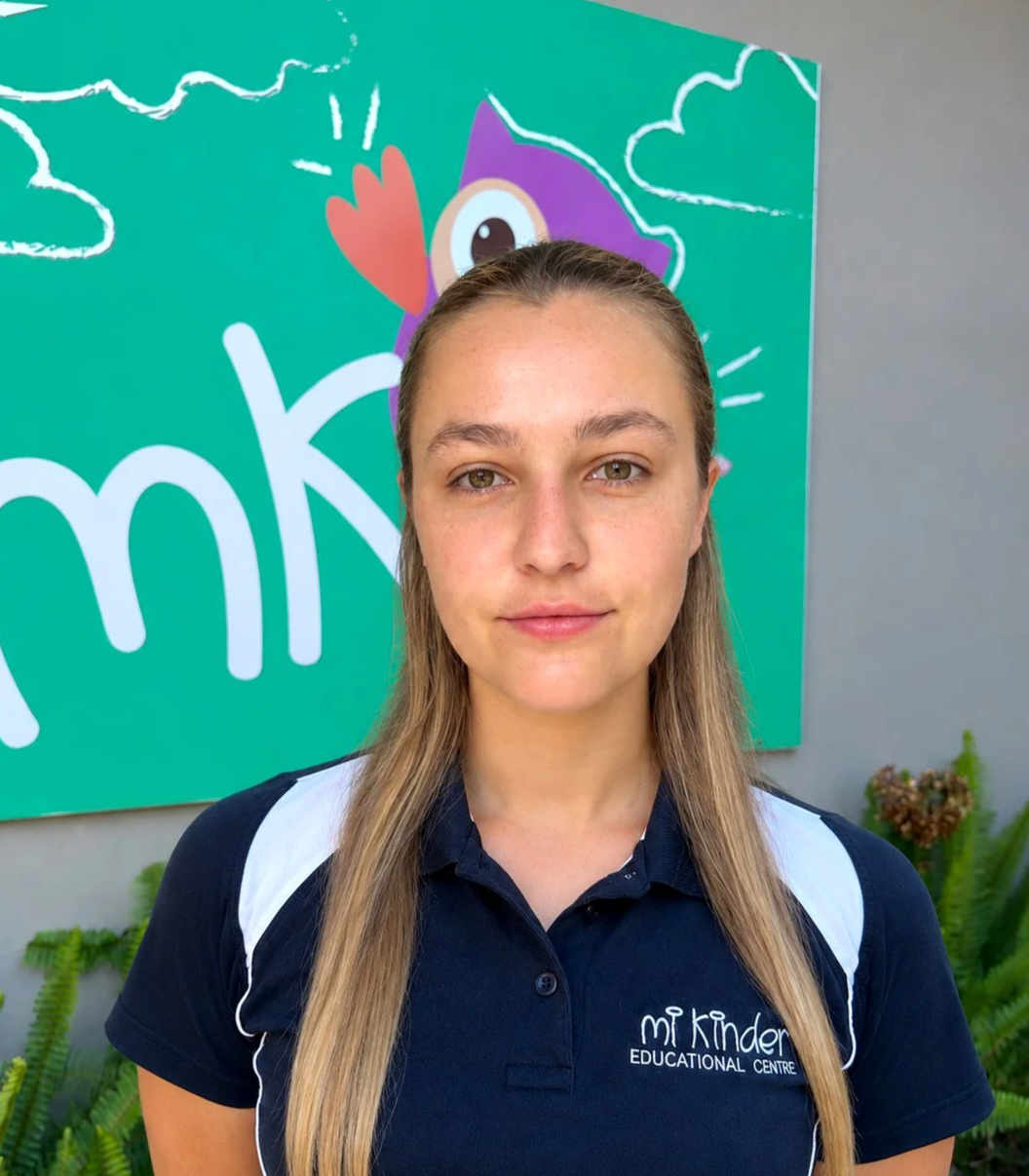 A young woman smiling, standing in front of a colorful owl mural at an educational center, wearing a navy blue polo shirt with white accents.