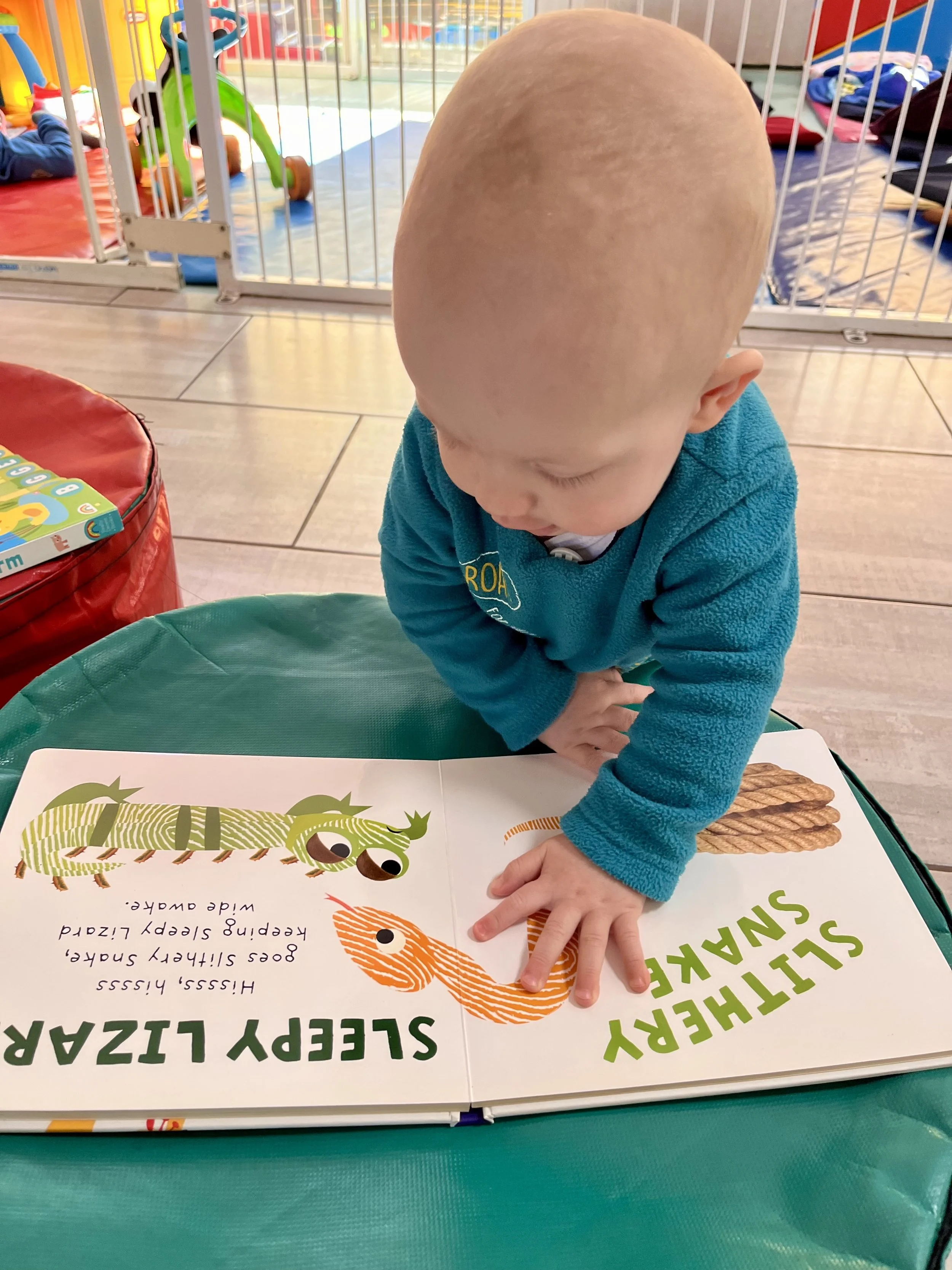 A young child with a bald head wearing a turquoise jacket, leaning on a table and pointing at a colorful children's book with illustrations of a caterpillar and a snake.