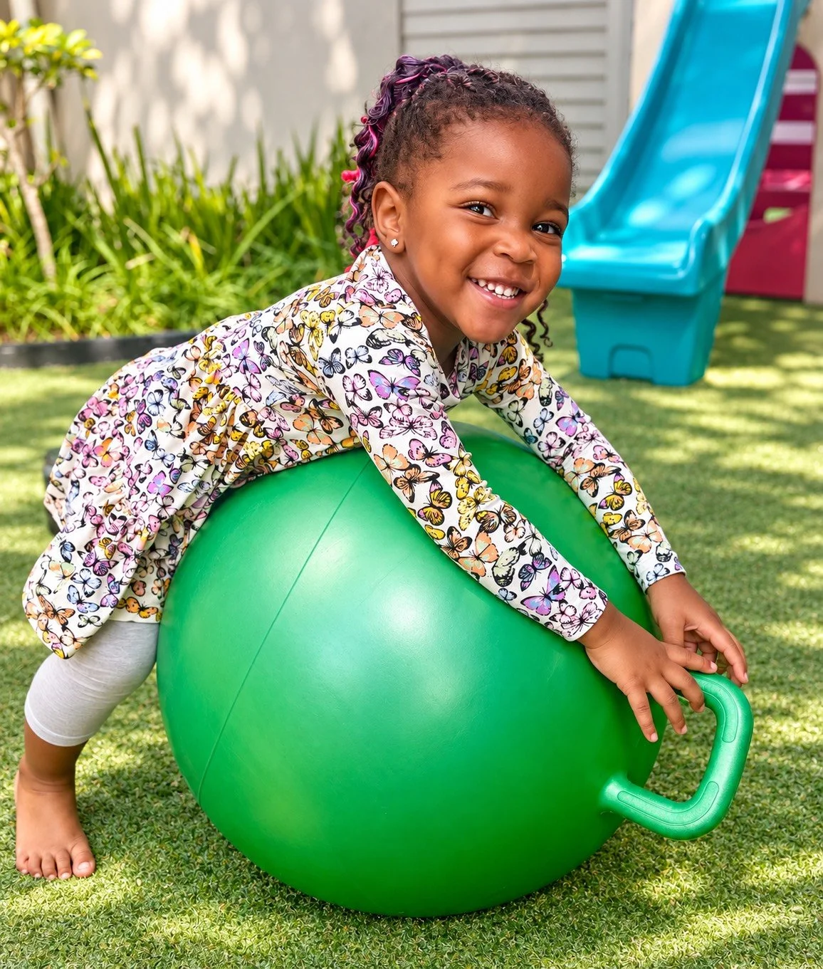 A smiling young girl with colorful braids playing on a large green exercise ball in a backyard, with a blue slide and greenery in the background.