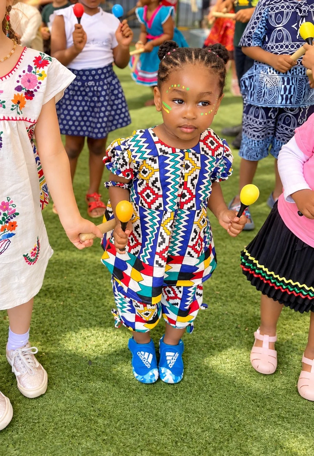 A young girl dressed in a vibrant, patterned outfit holding a maraca, standing among other children at an outdoor cultural event or festival. The children are wearing colorful clothing and face paint, and they are holding maracas.