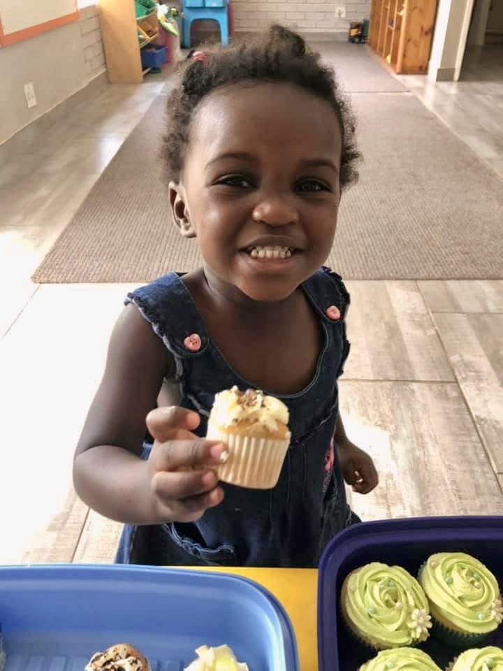 A young girl with dark skin and curly hair holding a cupcake with white frosting and sprinkles, smiling and standing in a room with colorful toys and wooden furniture.