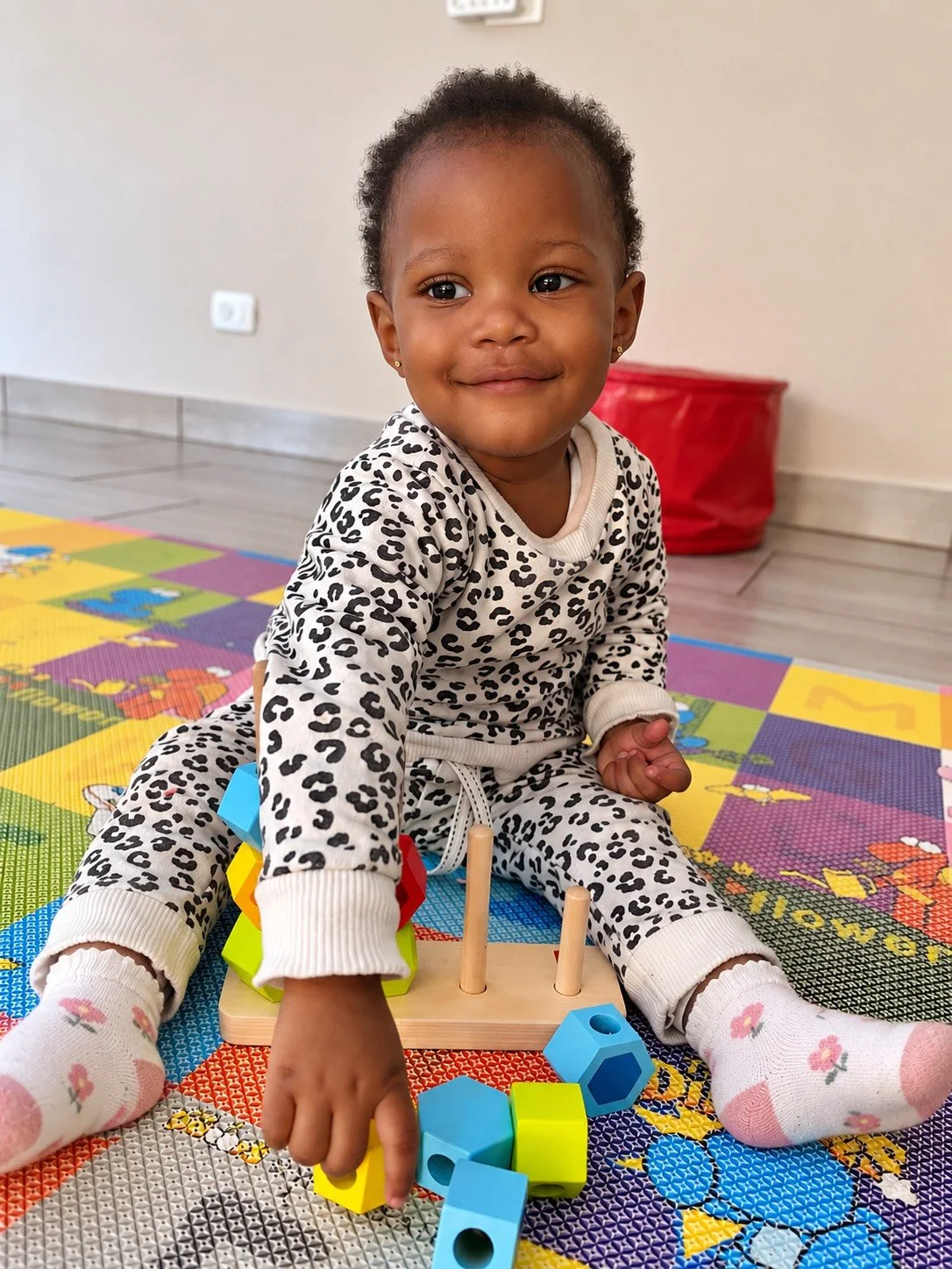 A young girl sitting on a colorful foam play mat, wearing a leopard print outfit, playing with building blocks, with a red storage bin in the background.