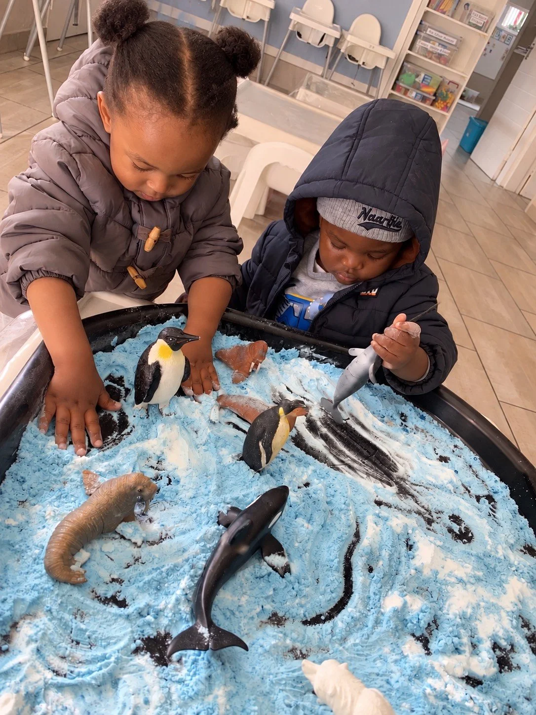 Two children playing with toy sea animals in a sandbox at an indoor play area.