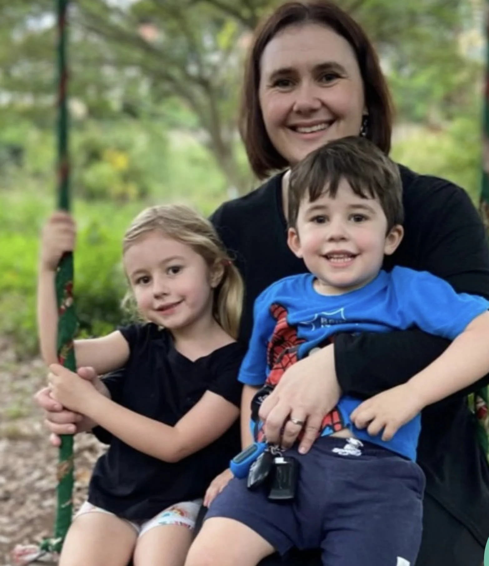 A woman with dark brown hair smiling, holding two young children on her lap outdoors on a swing set, green trees in the background.