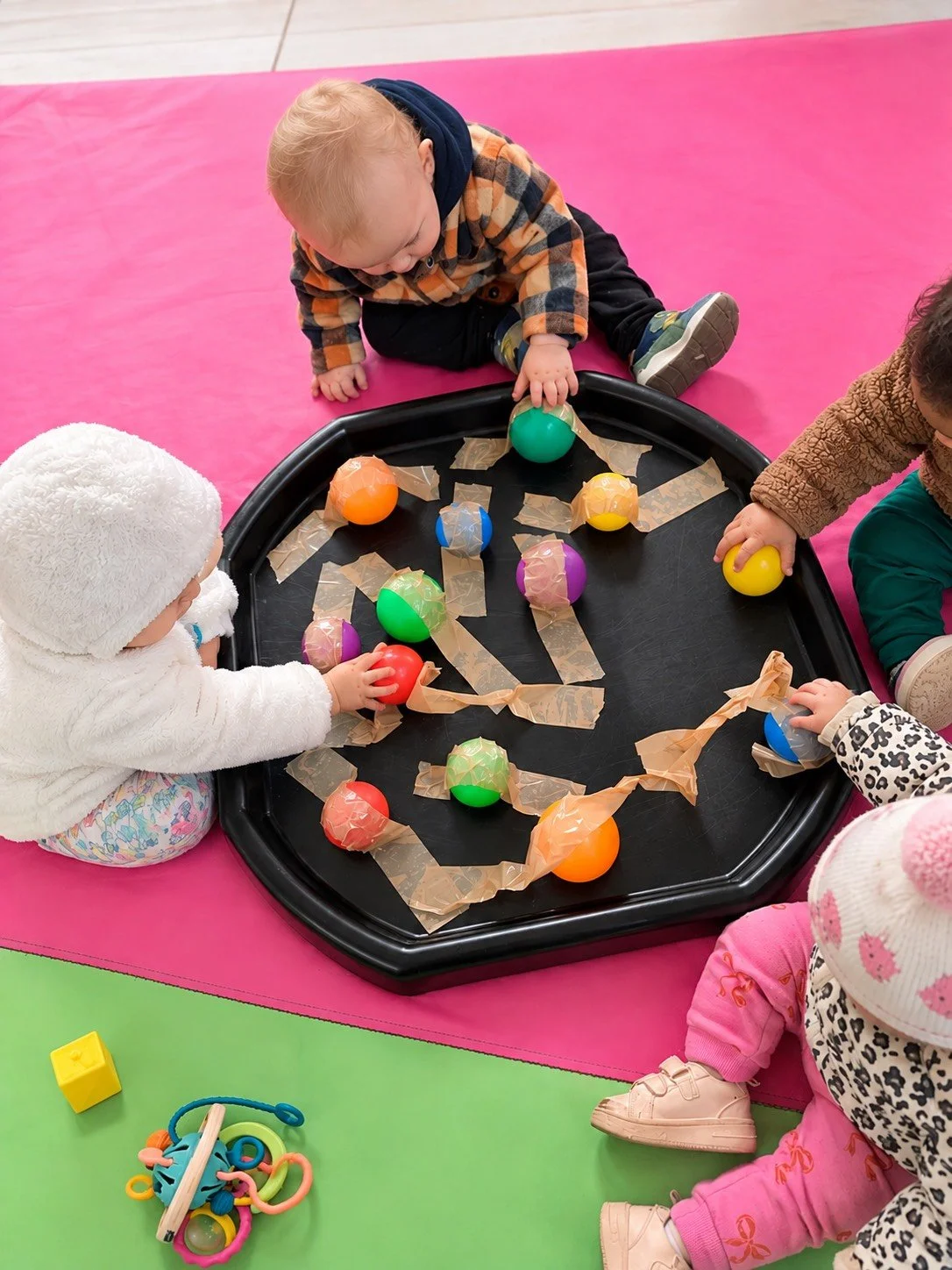 Children playing with colorful balls on a black tray, surrounded by pink and green mats, in a daycare or playroom setting.
