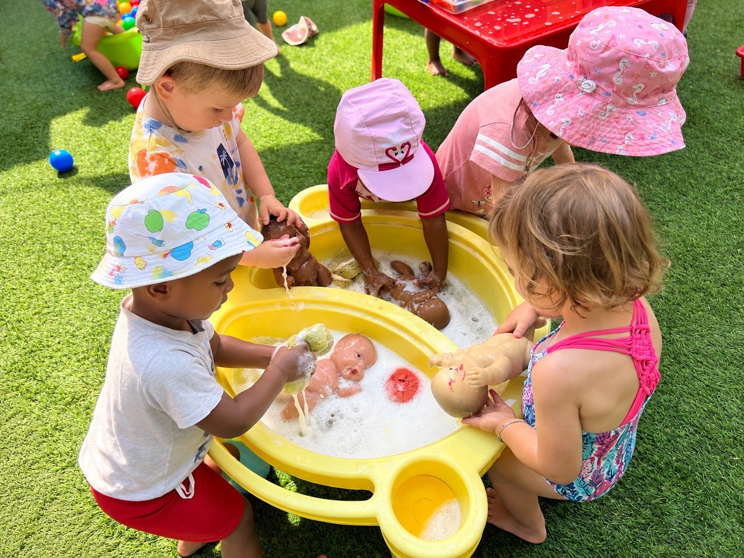 Children playing with water and toys in small yellow plastic pools outdoors, wearing colorful hats and swimsuits.
