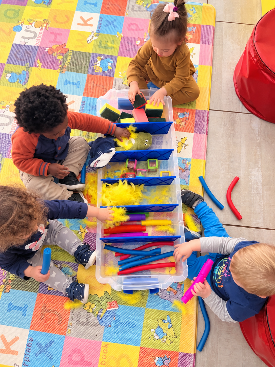 Children playing with colorful foam tubes and toys on a multicolored alphabet-themed play mat.