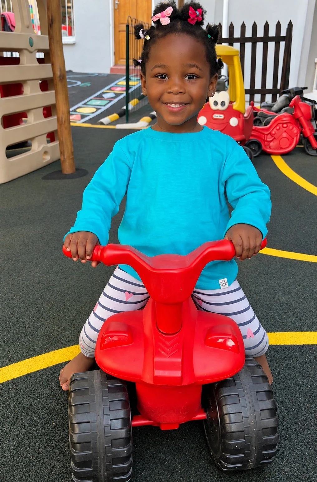 A young girl with braided hair and pink bows smiling while sitting on a red toy ride-on motorcycle in a playground with colorful play equipment and a black fence in the background.