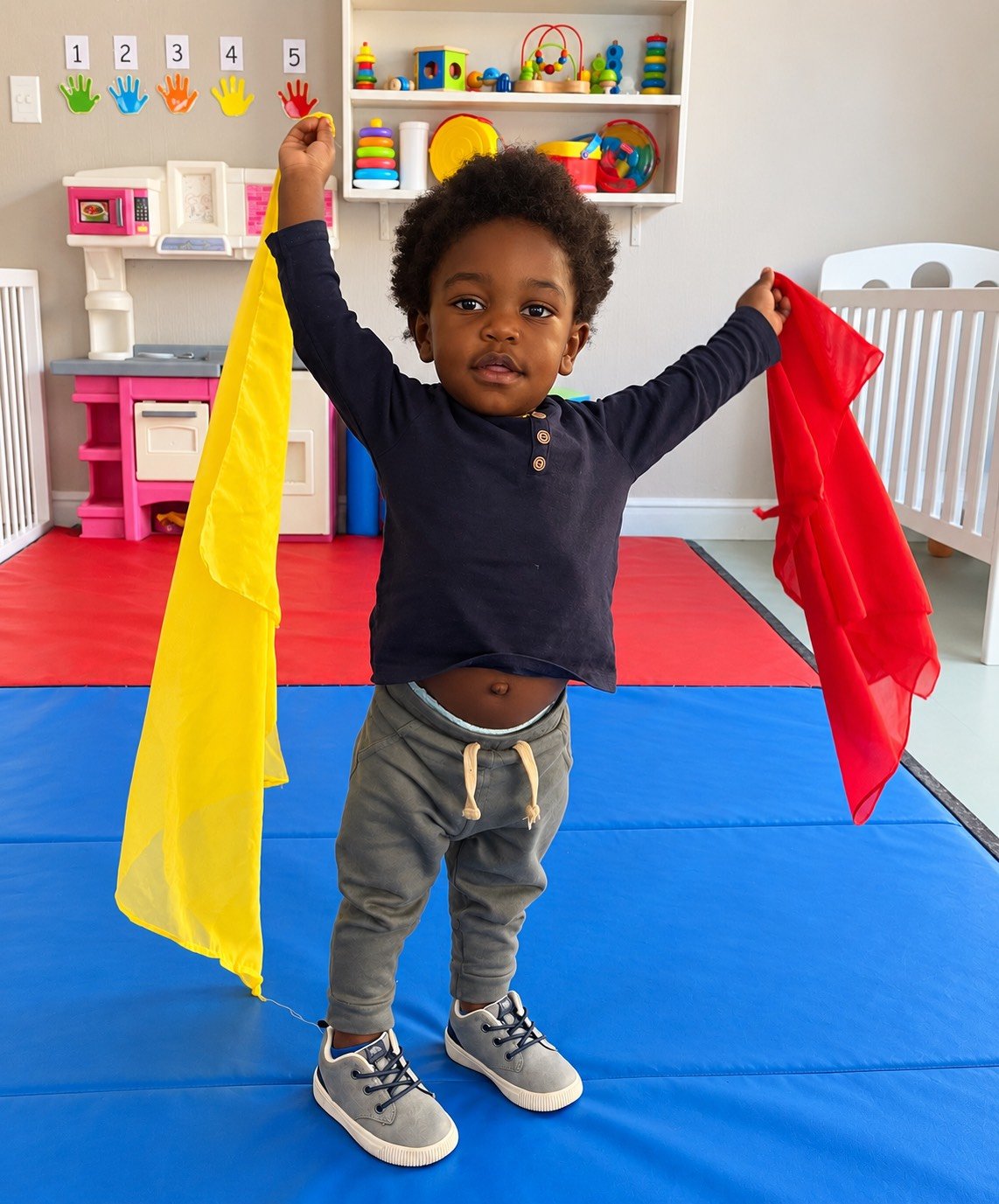 Young child standing on a blue mat in a playroom holding yellow and red cloths with his arms raised.
