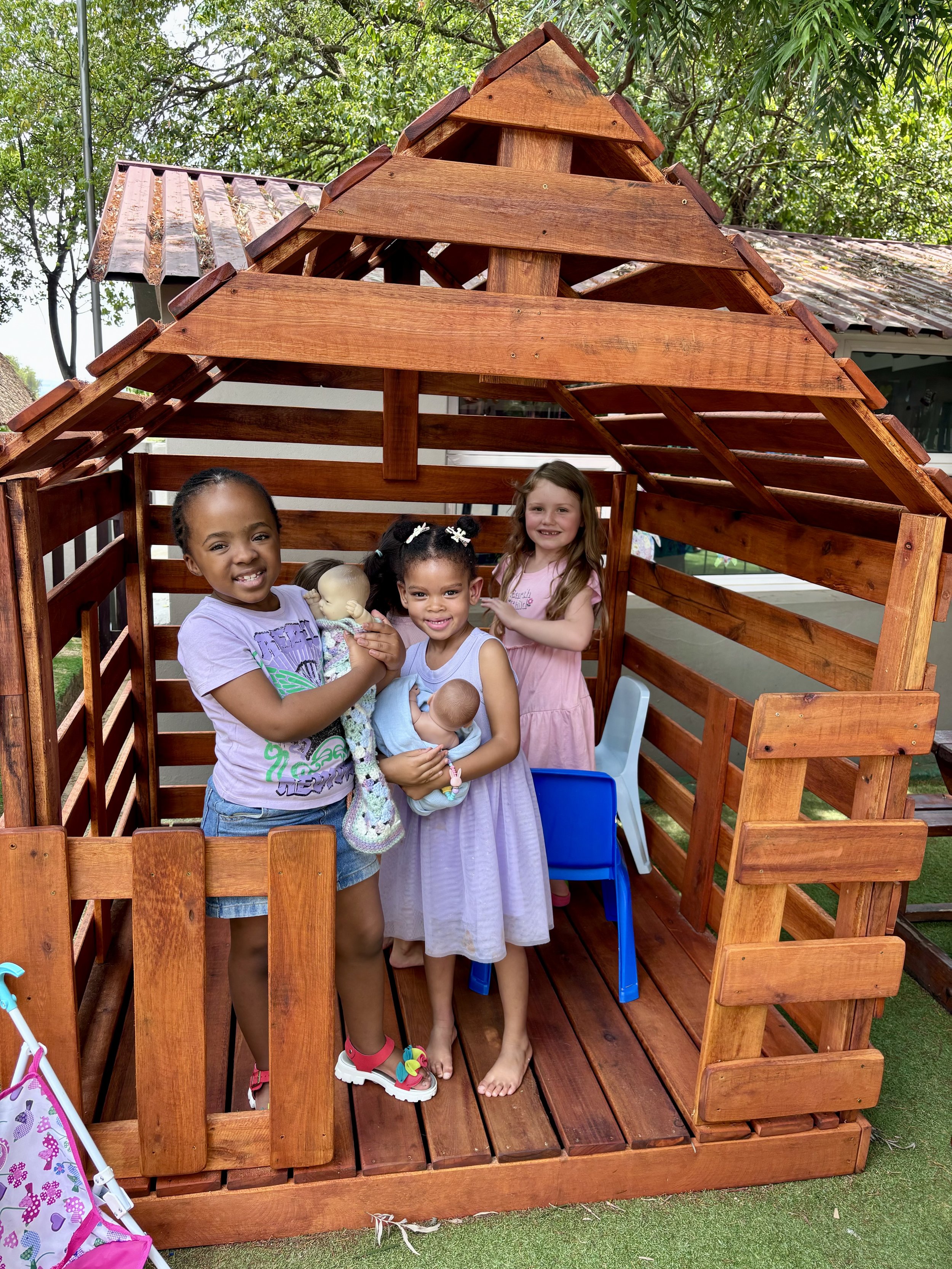 Four young girls standing inside a wooden playhouse outdoors, with two girls holding dolls.