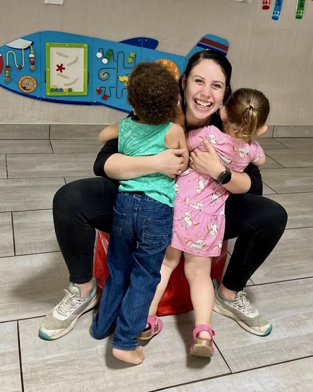 A woman with dark hair smiling and hugging two young children, one boy and one girl, in front of a colorful wall with children's artwork and a clock. The boy has curly hair and is wearing a green shirt and jeans, while the girl has light hair in pigt
