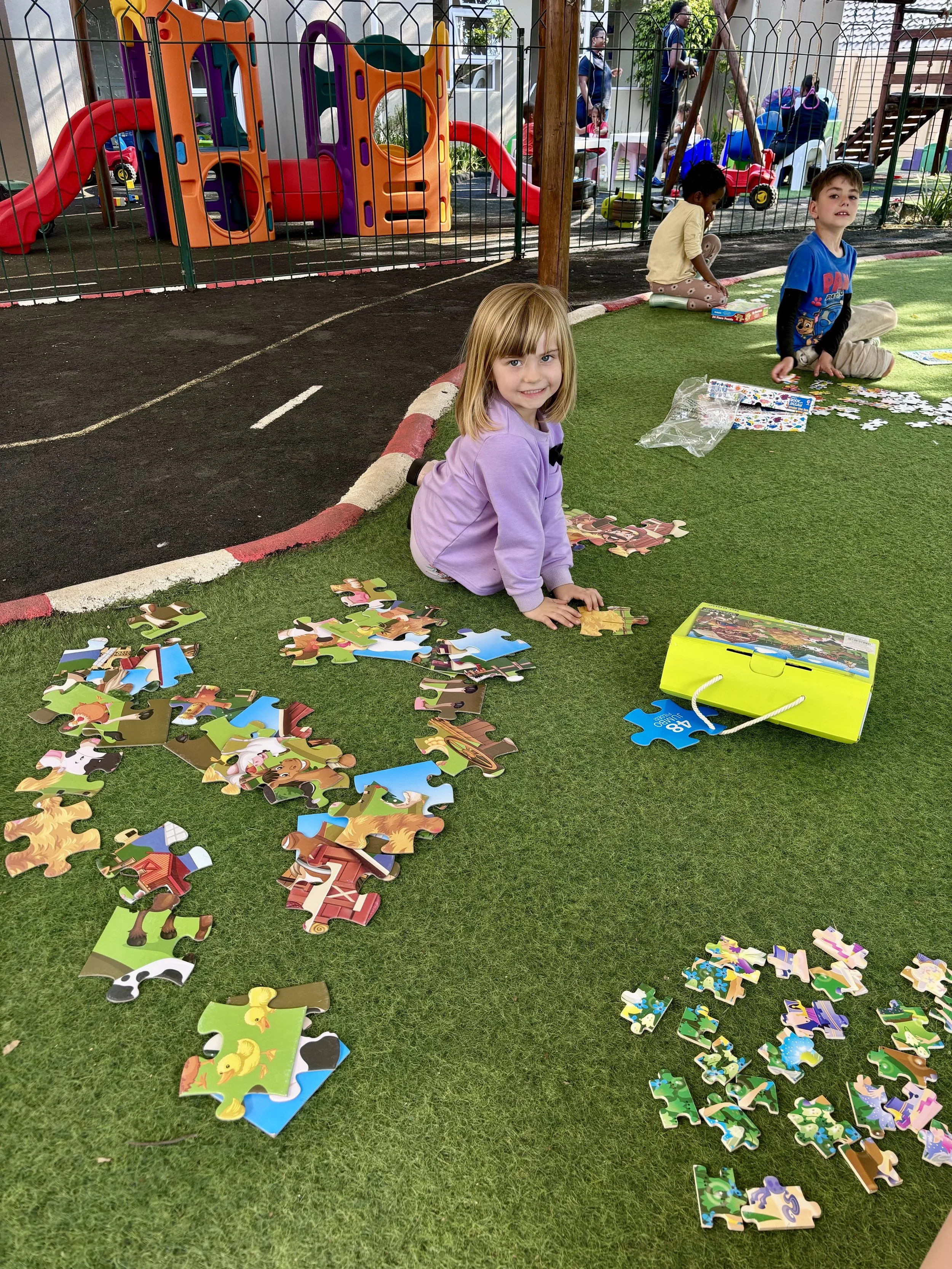 Children sitting on the grass playing with a jigsaw puzzle, with a playground visible behind them enclosed by a metal fence.