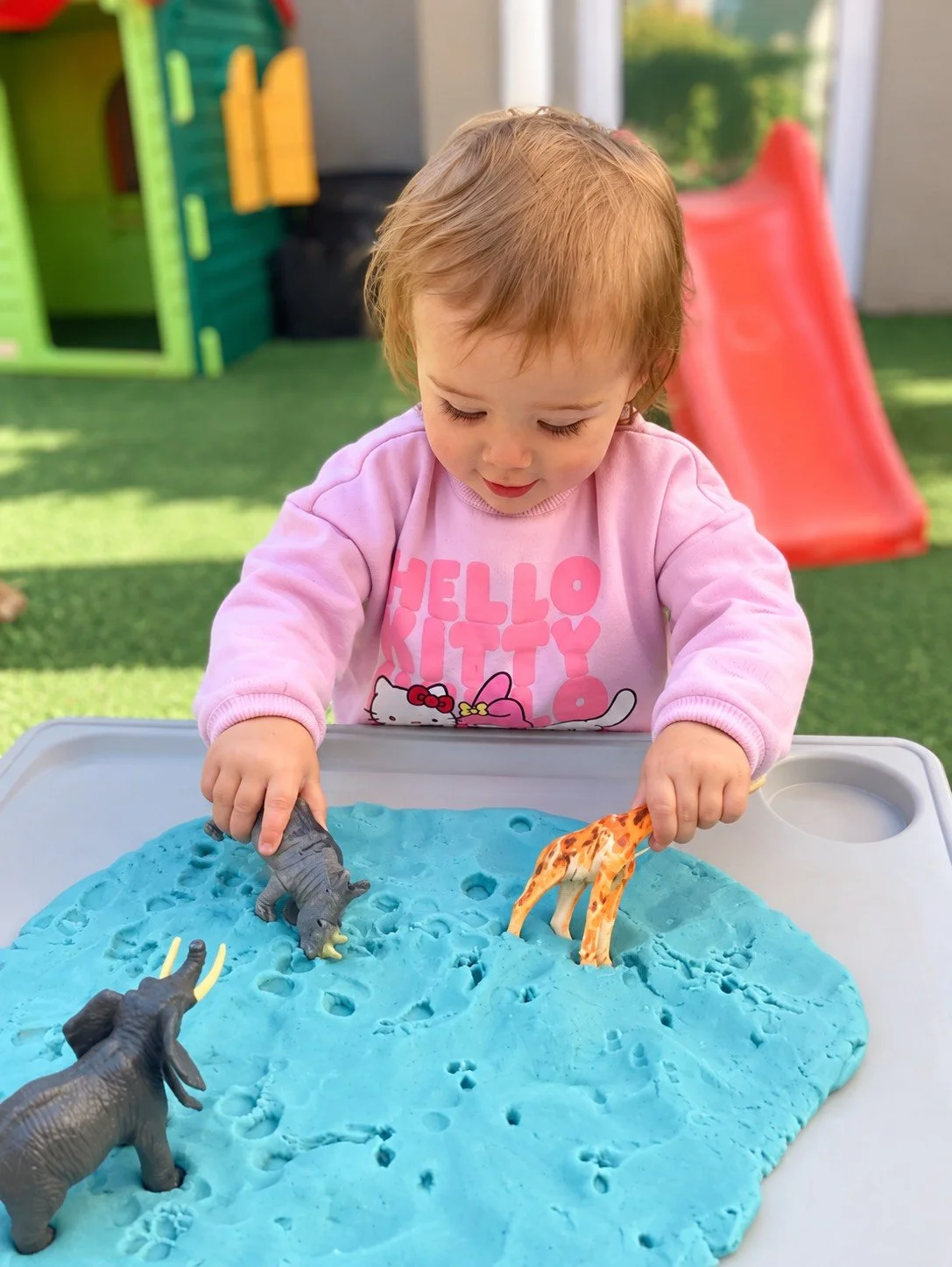 A young girl with light brown hair wearing a pink Hello Kitty sweatshirt plays with toy animals in blue modeling clay on a table outdoors. There is a red slide and playhouse in the background.