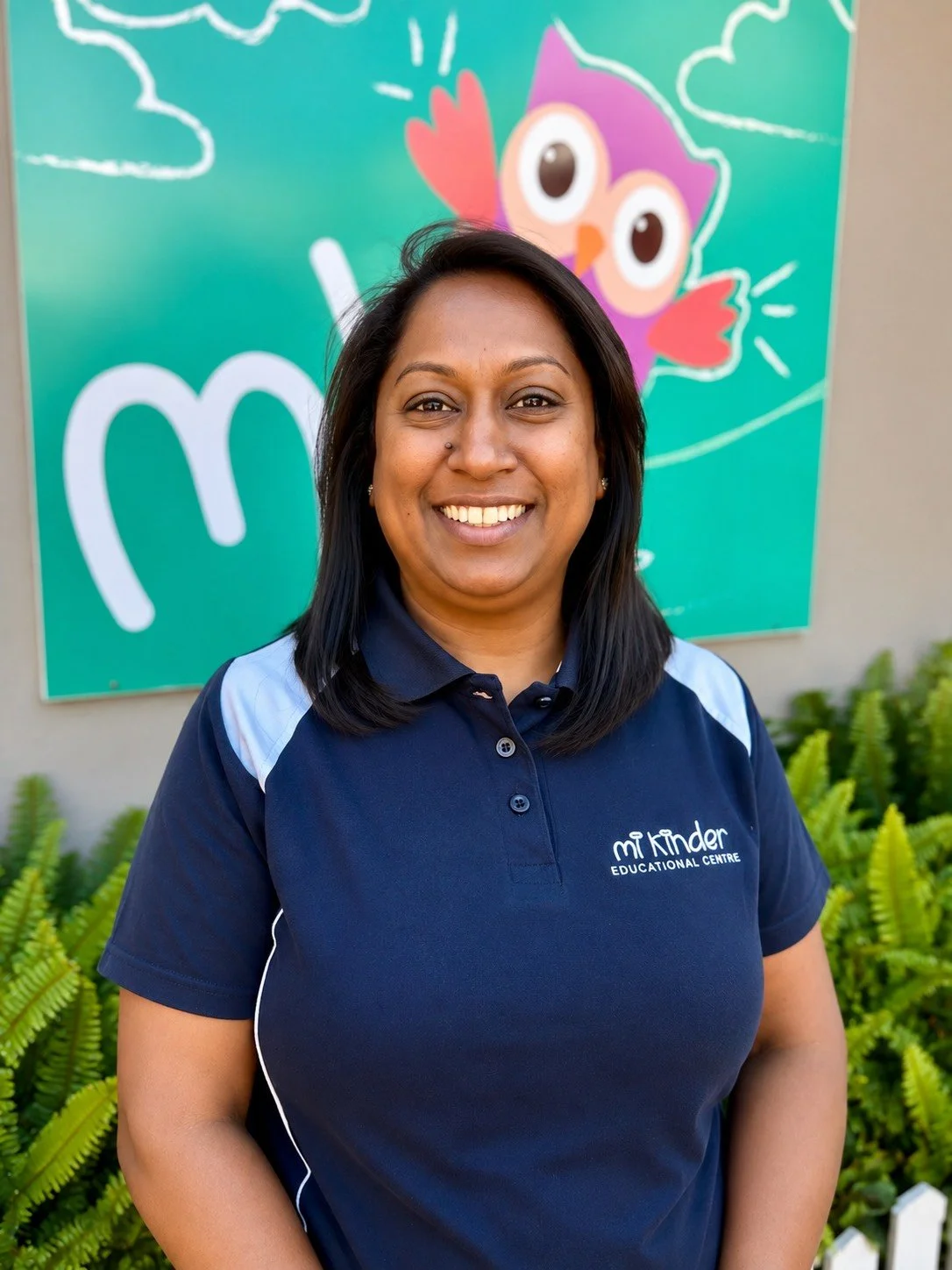 Smiling woman in navy blue polo shirt with 'Mi Kinder Educational Centre' logo standing outdoors in front of a colorful owl-themed sign and green plants.