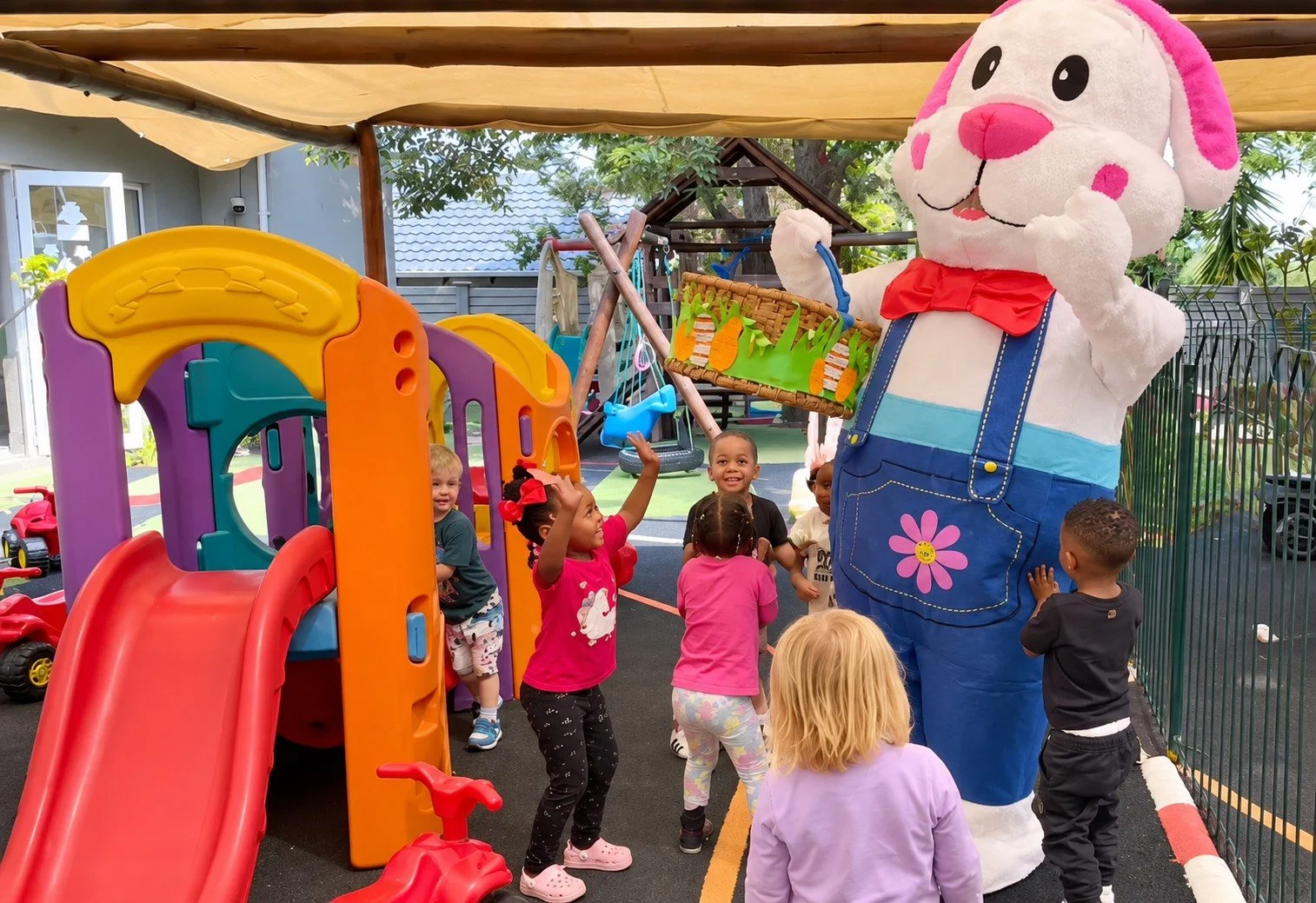 Children playing in a playground with a person in a large bunny costume holding a branch with carrots. The children are reaching towards the bunny and smiling. There is a colorful slide and swings in the background on a sunny day.