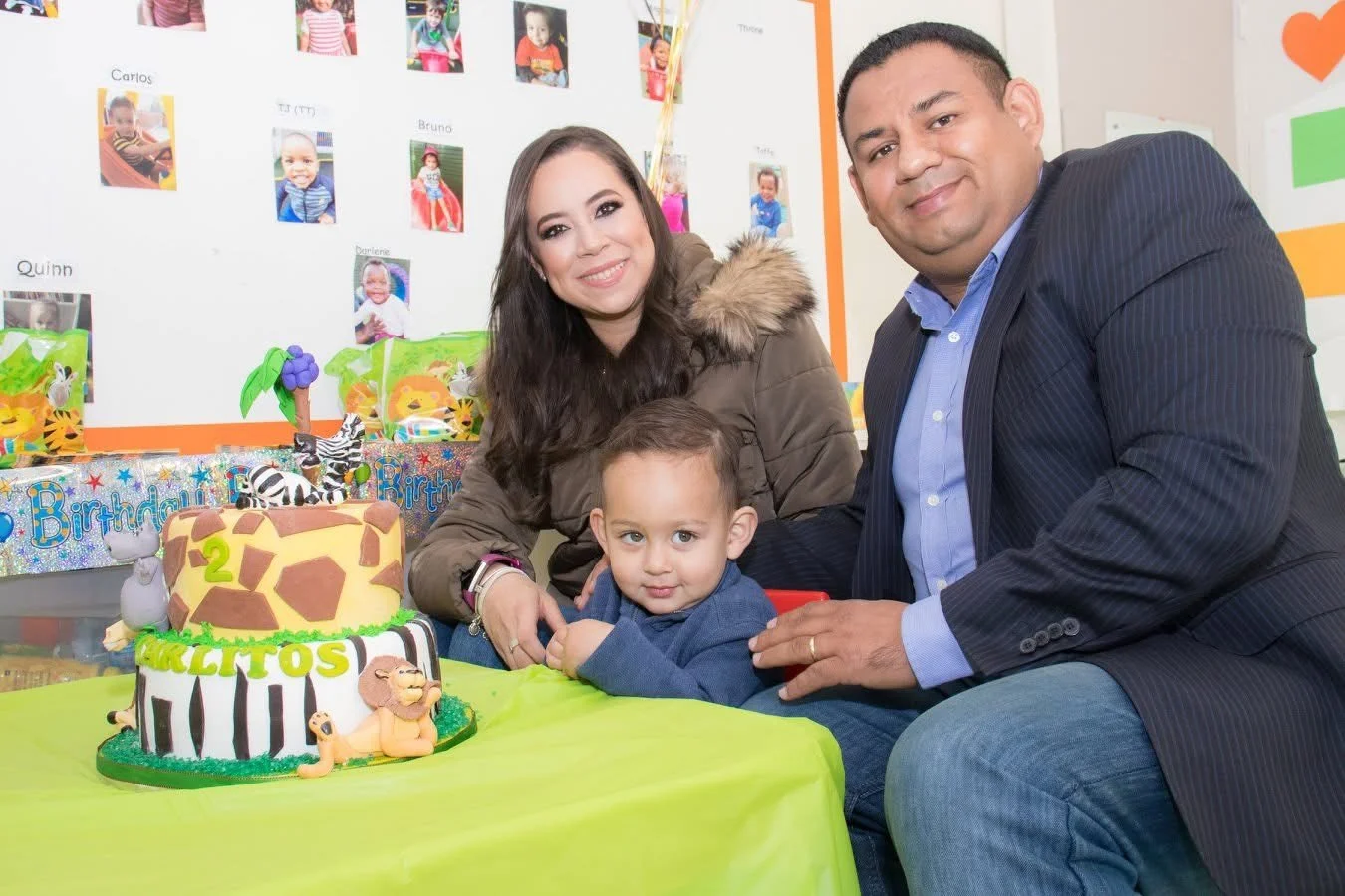 Family celebrating a child's birthday with a jungle-themed cake featuring a lion, giraffe, zebra, and hippo, at a party with colorful decorations and a display of children's photos in the background.