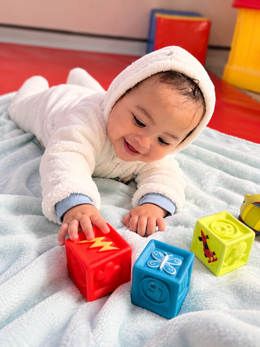 A baby in a white hooded jacket lying on a soft light blue blanket, playing with colorful plastic toy blocks shaped like a lightning bolt, a butterfly, and a creature. The baby is smiling and focused on the blocks.