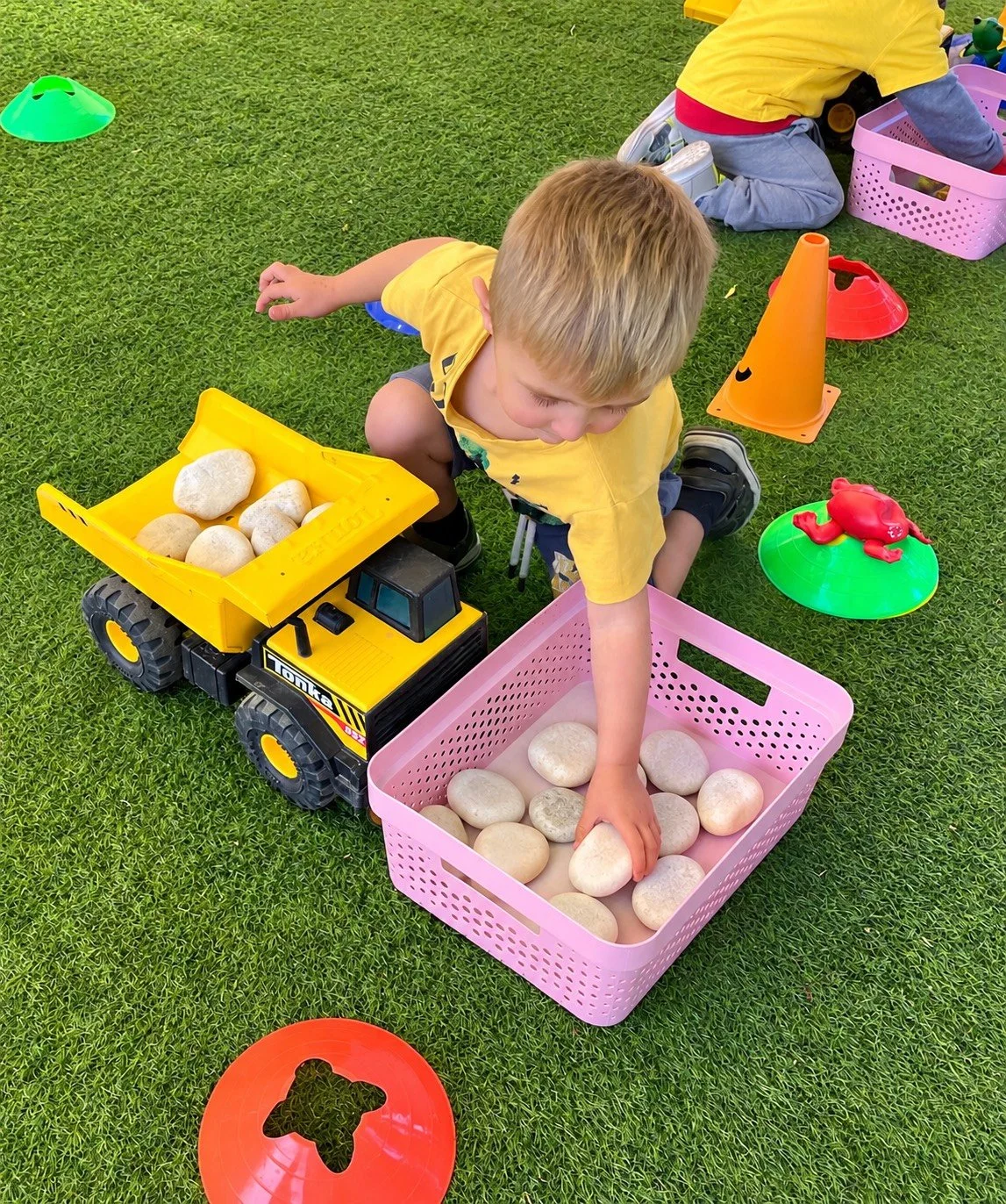 Young boy playing outdoors with toy rocks and construction truck, surrounded by colorful cones and toys on a grassy area.