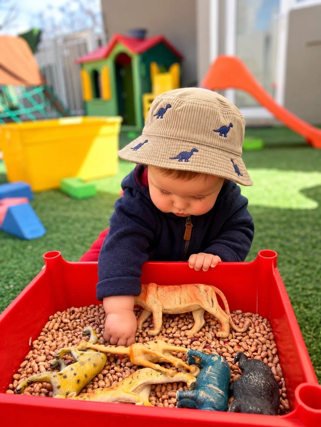 A young child wearing a beige hat with dinosaur prints and a navy jacket plays in a red sandbox filled with toy dinosaur figurines and small pebbles. In the background, there's a colorful playhouse, slide, and sandbox on a grassy outdoor play area.