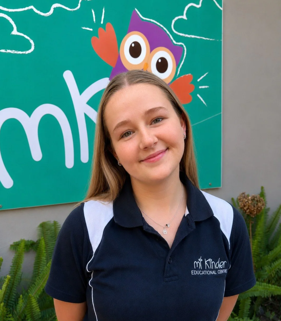 A young woman in a navy and white polo shirt with 'mt Kinder Educational Centre' logo, standing in front of a colorful cartoon owl and a 'mko' sign, with greenery around her.