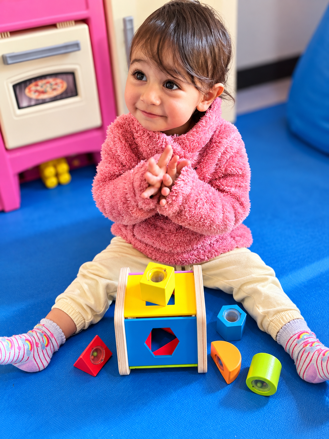 A young girl with brown hair and big eyes, wearing a pink fuzzy jacket, cream pants, and striped socks, sitting on a blue floor, playing with colorful wooden shape sorter toys.