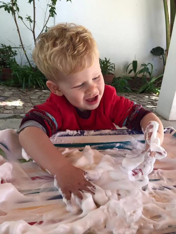 Young boy with curly blonde hair, wearing a red shirt, playing with foam or shaving cream on a table outdoors.