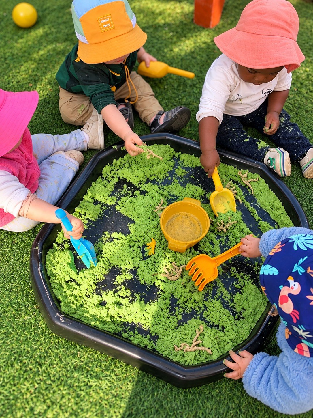 Children playing with toy dinosaurs in a sandbox with green textured material, wearing colorful hats and outdoor clothing, on a grassy area.