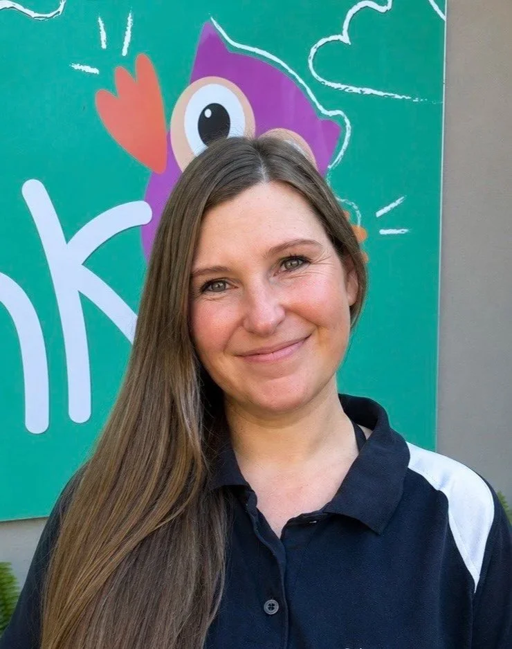 A woman with long brown hair smiling outdoors, standing in front of a colorful cartoon mural featuring a purple creature with big eyes, a heart shape, and abstract designs.