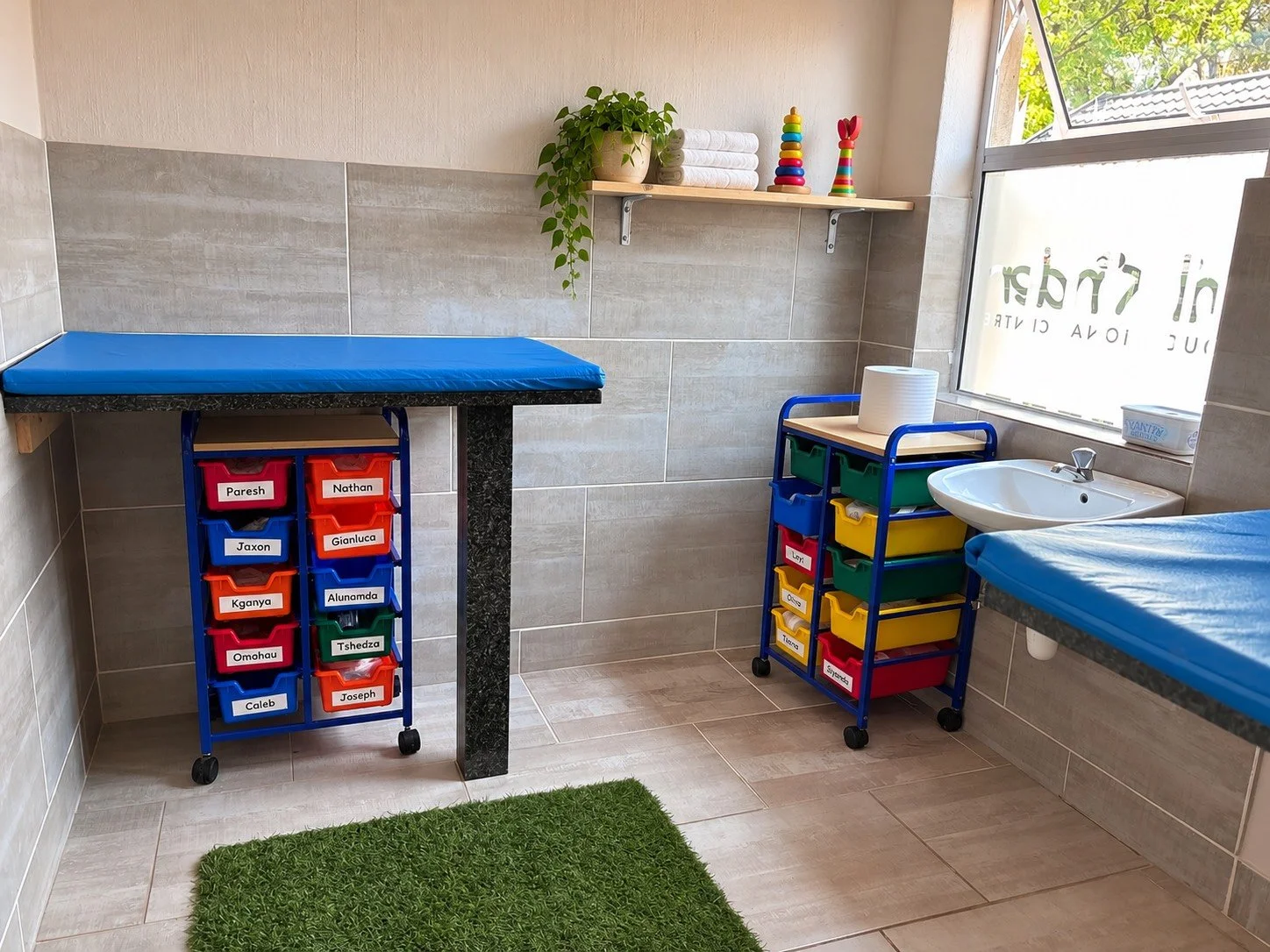 A colorful classroom or daycare room with labeled storage bins and a changing table, a sink, and a window with a view of trees outside.