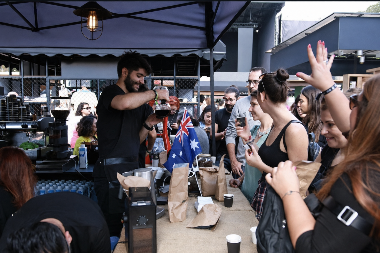 A barista pouring coffee for customers at an outdoor coffee stand, Event, expo con and a crowd gathered around.