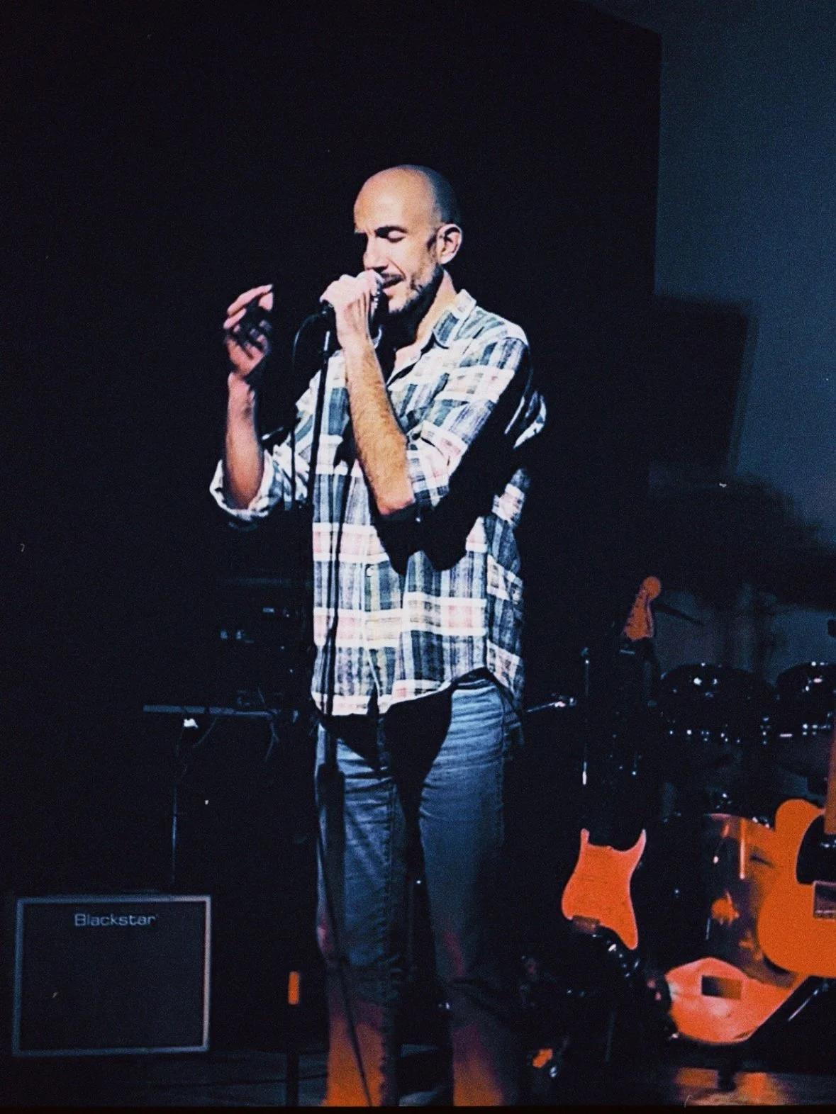 A man with a shaved head and beard singing into a microphone on stage, wearing a plaid shirt, with musical instruments and amplifier in the background.
