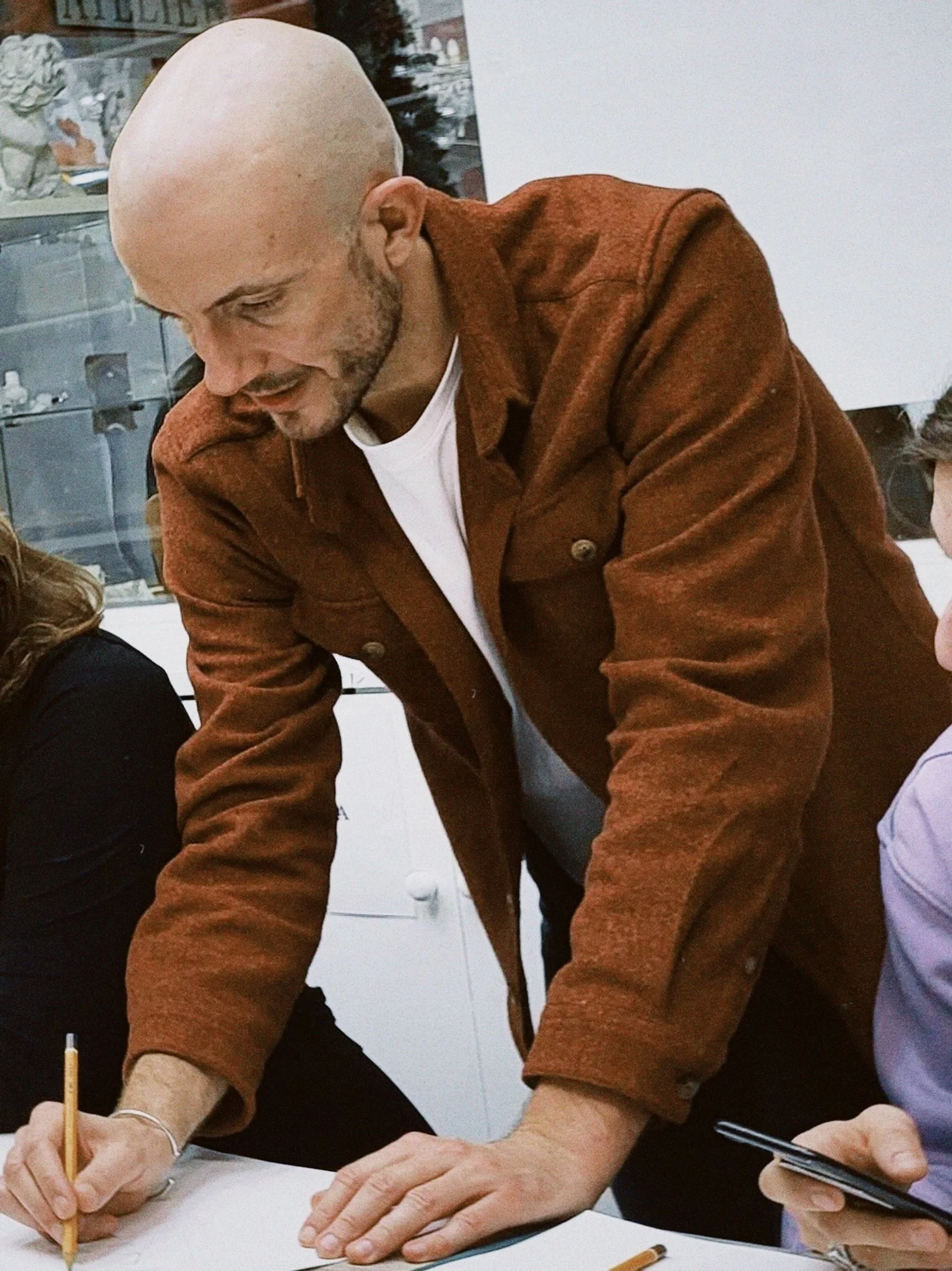 Close-up of a bald man with a beard wearing a brown jacket, writing with a pencil on paper at a table.
