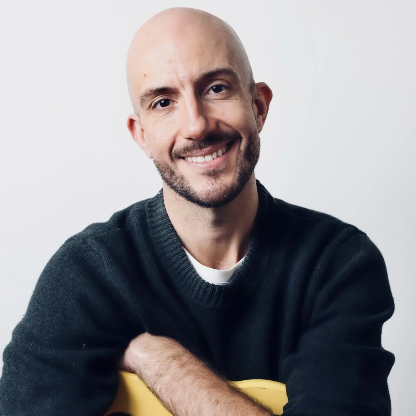 A smiling bald man with a beard wearing a dark sweater, with his arms crossed, holding a yellow object, in front of a plain white background.