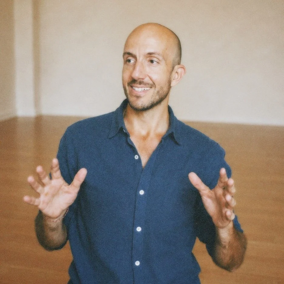 A bald man wearing a dark blue button-up shirt smiling and gesturing with his hands, indoors with a wooden floor and light-colored wall.