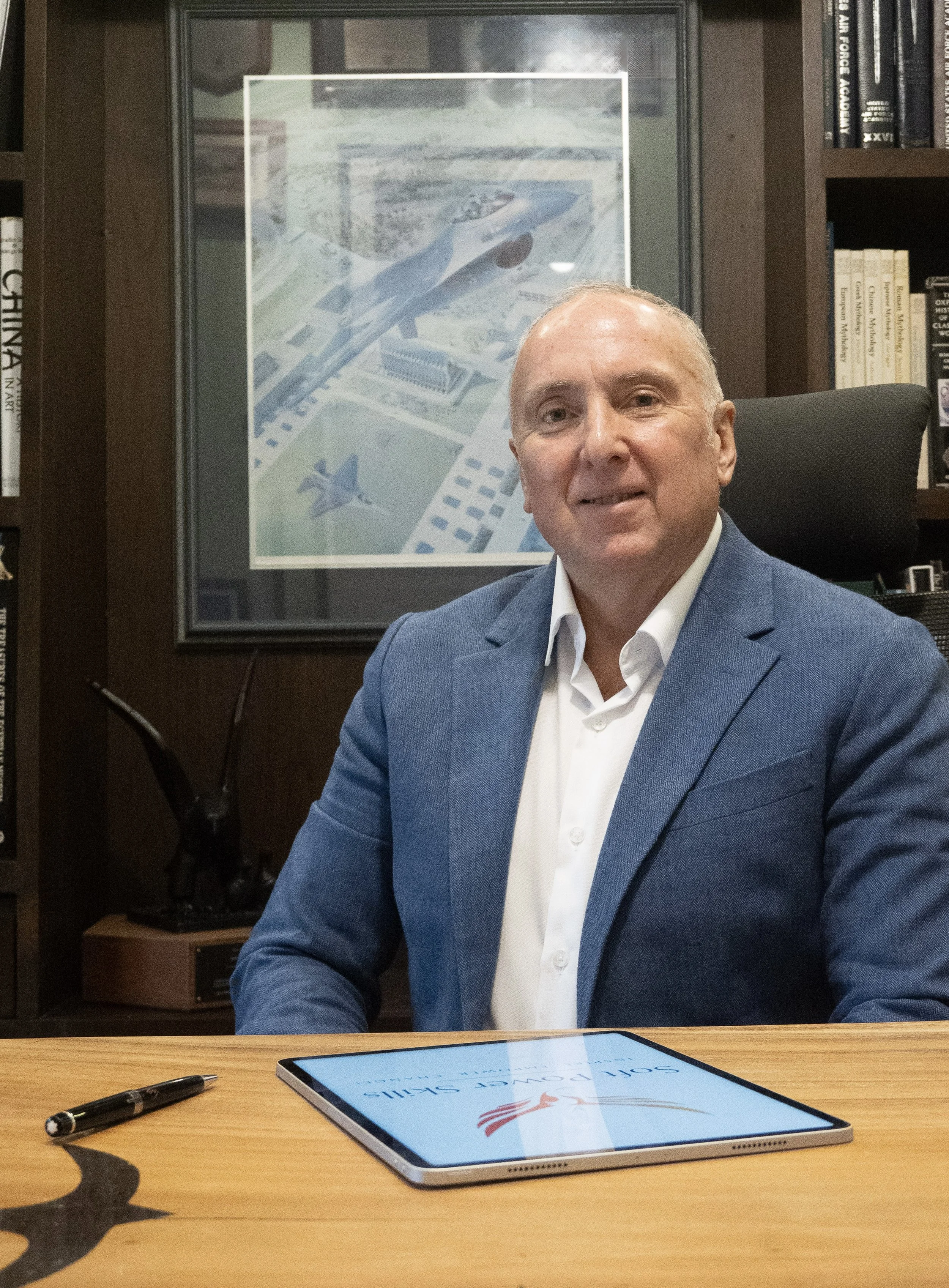 A man in a blue suit sits at a table in an office. In front of him on the table is a tablet displaying a sign with the words 'SOAR WITH US!' and a red, white, and blue logo. There is a black marker and a pen on the table. Behind the man, there is a framed aerial photo of an airport with a plane and structures, and bookshelves filled with books.