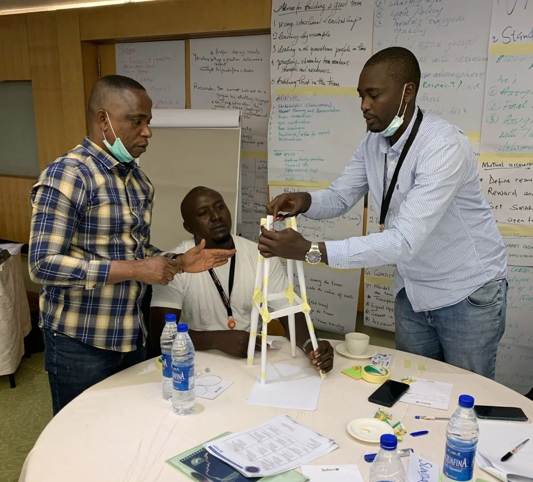 Three workshop participants collaborate on a straw-and-tape structure at a table, with handwritten flip-chart notes behind them.