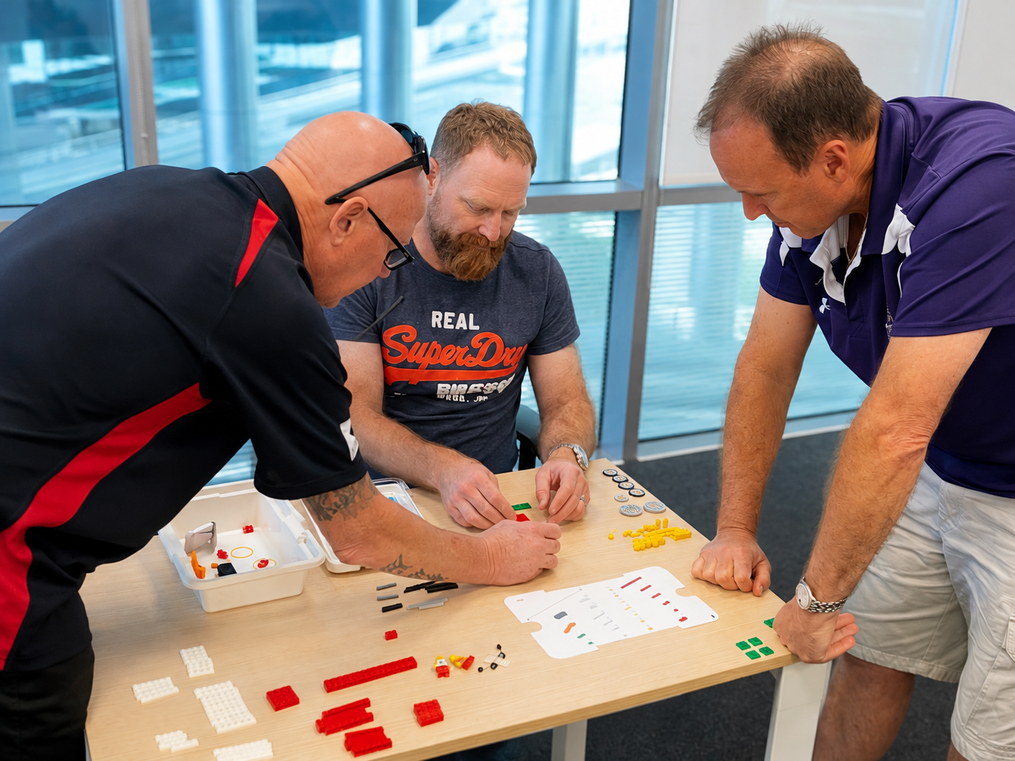 Three men are working together at a table assembling pieces of a LEGO set. The table has various LEGO bricks and instructions. The men are leaning over the table, focused on their task, in a modern office or building with large windows behind them.