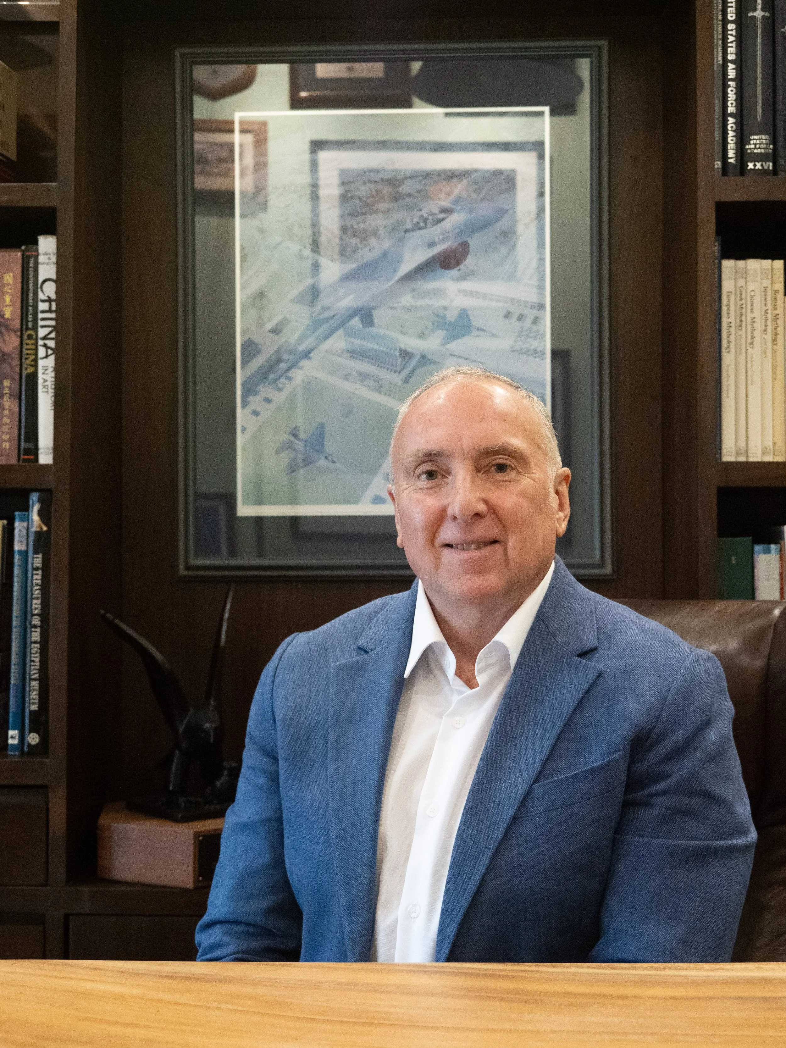A man in a blue suit sitting at a desk in front of a framed airplane poster and bookshelves.