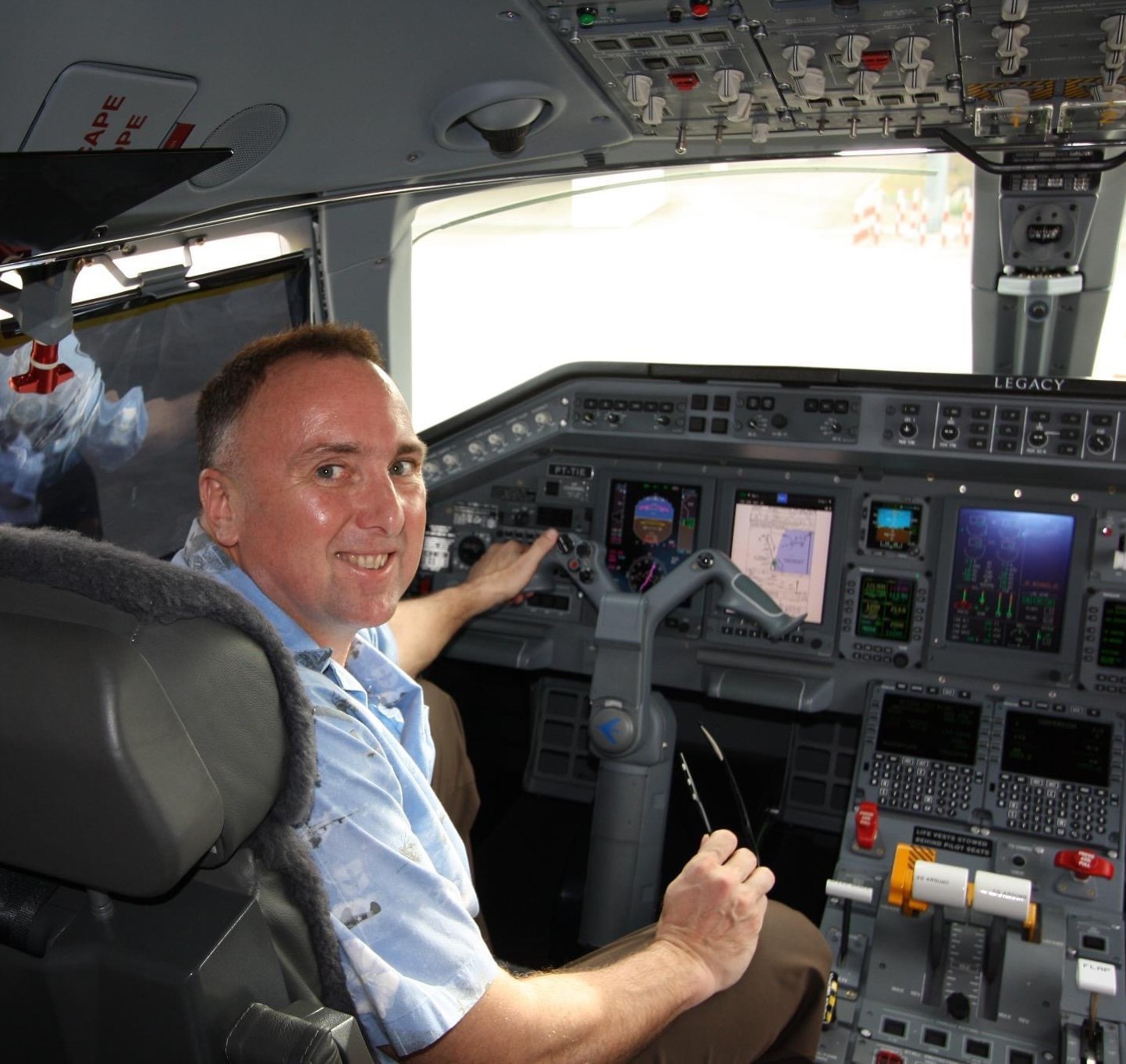 A smiling man sitting in the cockpit of an airplane, holding the control yoke, with various flight instruments and controls visible on the dashboard.