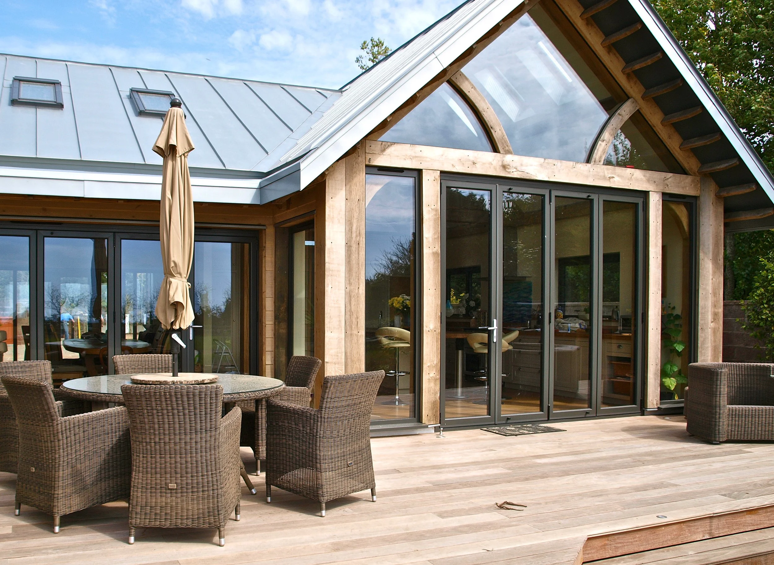 A modern house with large glass sliding doors, a wooden frame, and a metallic roof. There is an outdoor patio area with wicker chairs and a round table with an umbrella. The house has a curved window on the upper level and is surrounded by greenery under a partly cloudy sky.