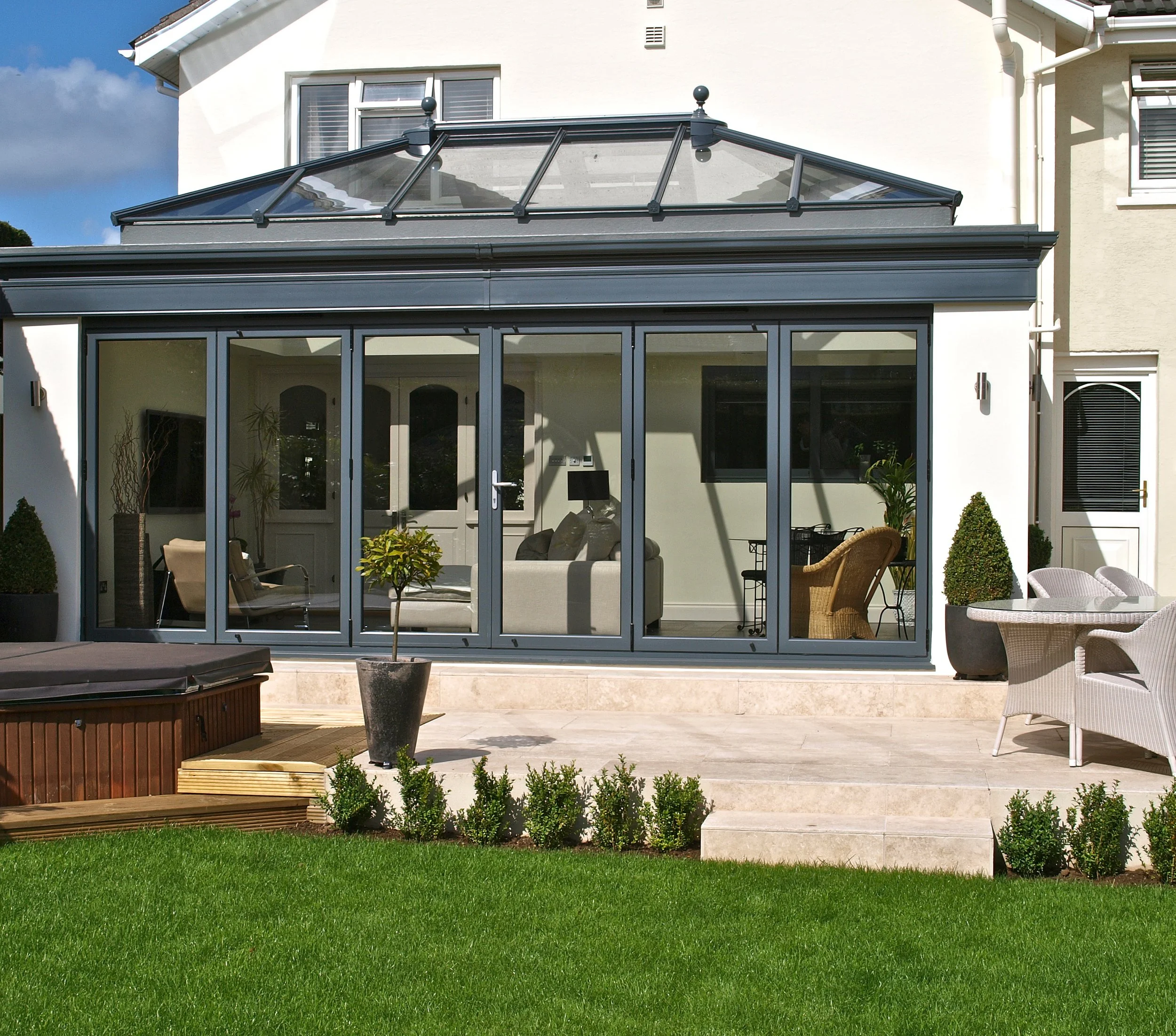 A modern residential house with a glass conservatory attached to the back yard. The conservatory has glass doors and windows, with patio furniture inside. There are small landscaped plants and a wooden fire pit on the patio, with a well-maintained green lawn in the foreground.