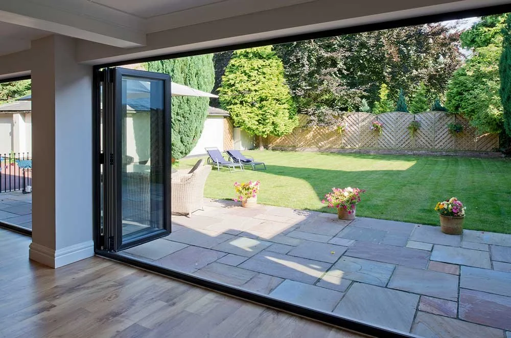 Open sliding glass door leading to a backyard garden with a lawn, potted flowers, trees, and outdoor lounge chairs.