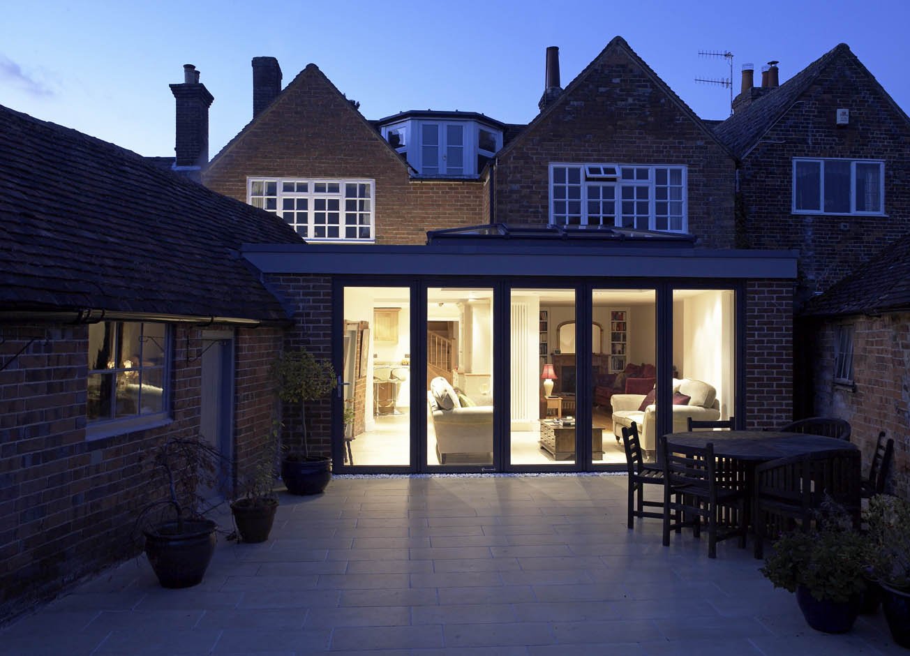 View of a backyard with a patio and glass-enclosed living space, illuminated by indoor lights at dusk, surrounded by brick houses.