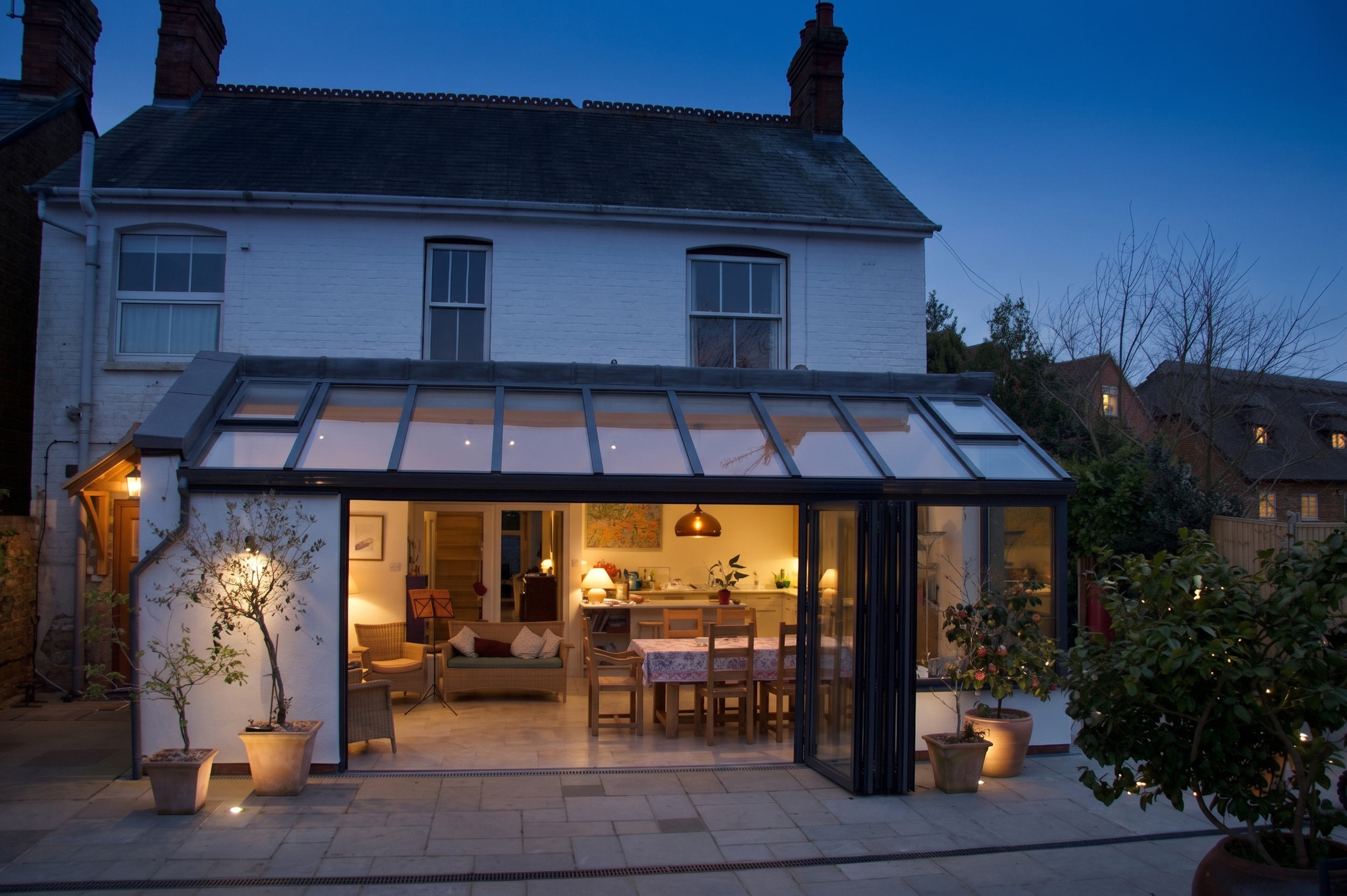A house with a glass-paned conservatory attached, illuminated from inside, with outdoor potted plants and trees, during evening twilight.