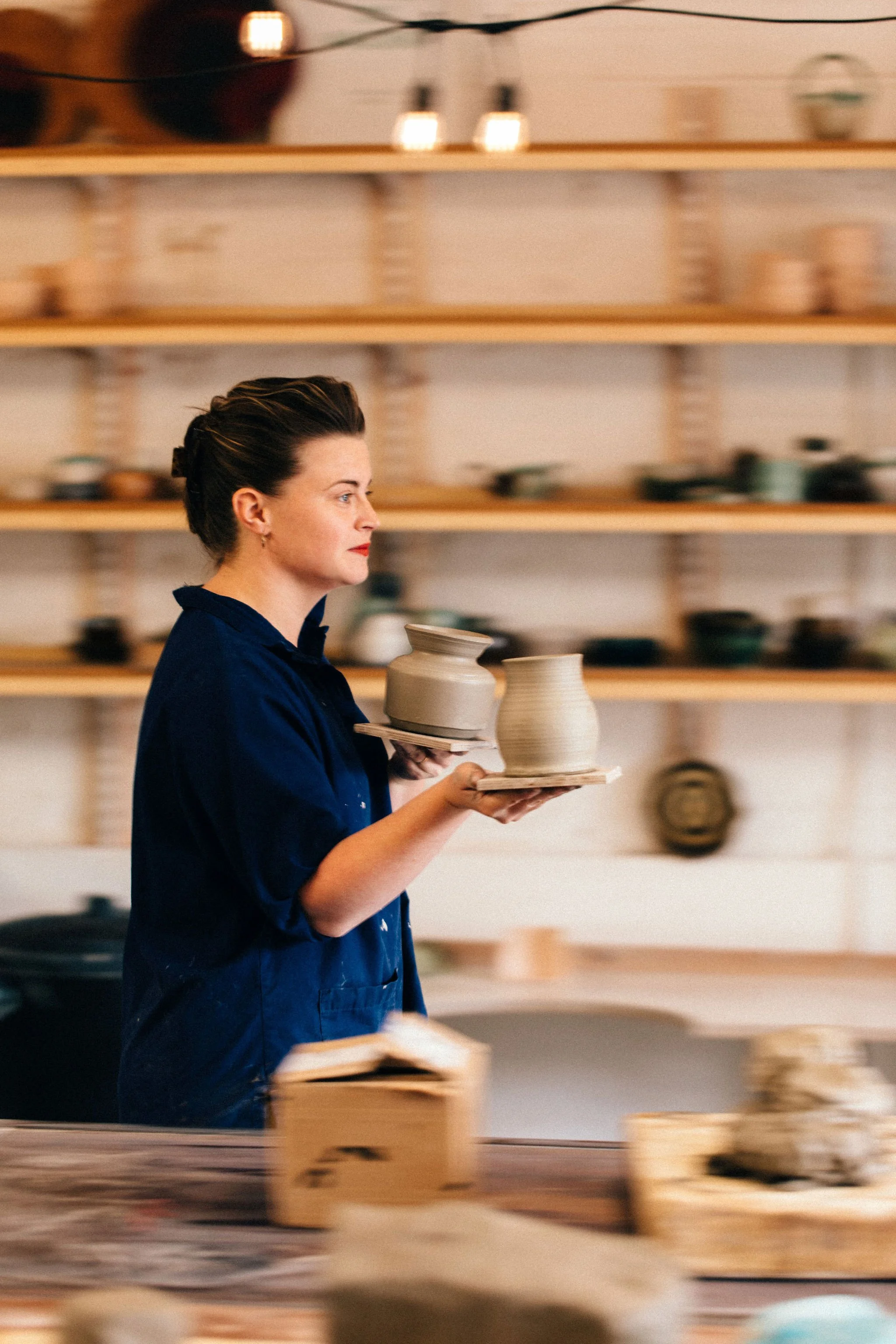 A woman with dark hair in a bun, wearing a dark blue shirt, is holding two pottery vases on a tray in a pottery studio. There are wooden shelves with pottery and ceramics behind her.