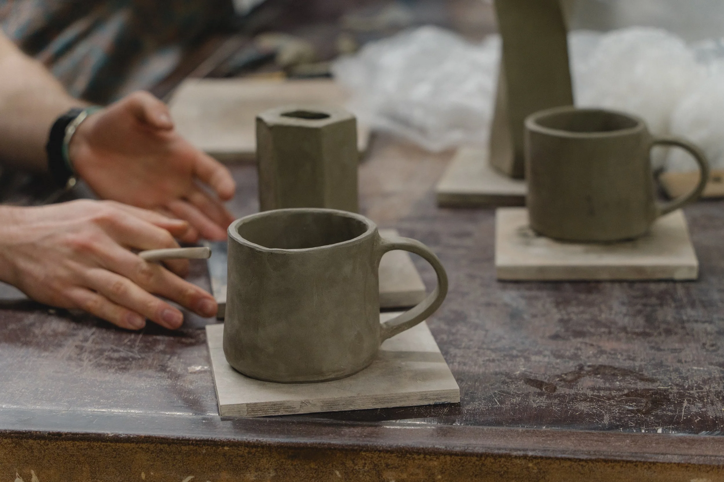 People working on pottery mugs, shaping and detailing them with tools on a worktable.