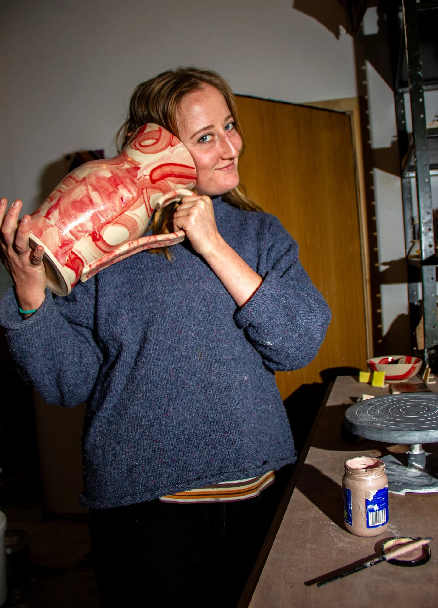 A smiling woman holding a decorative ceramic mug shaped like a frog at a craft or art studio, with painting supplies and a pottery wheel on the table.