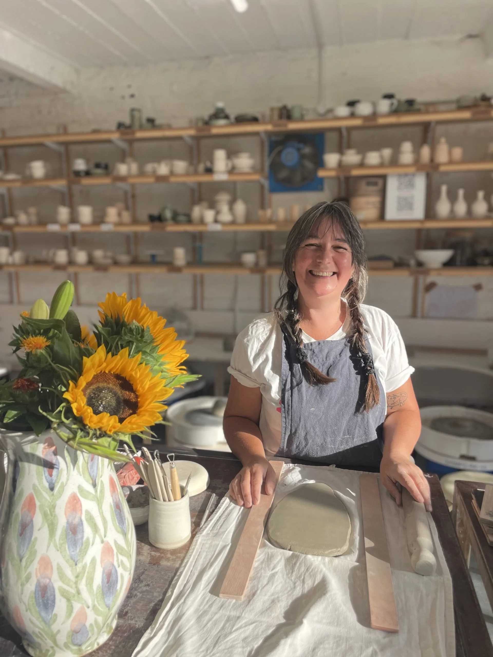 A woman smiling while working on a pottery wheel in a ceramics studio. There are shelves with various ceramics and a large sunflower bouquet in the foreground.